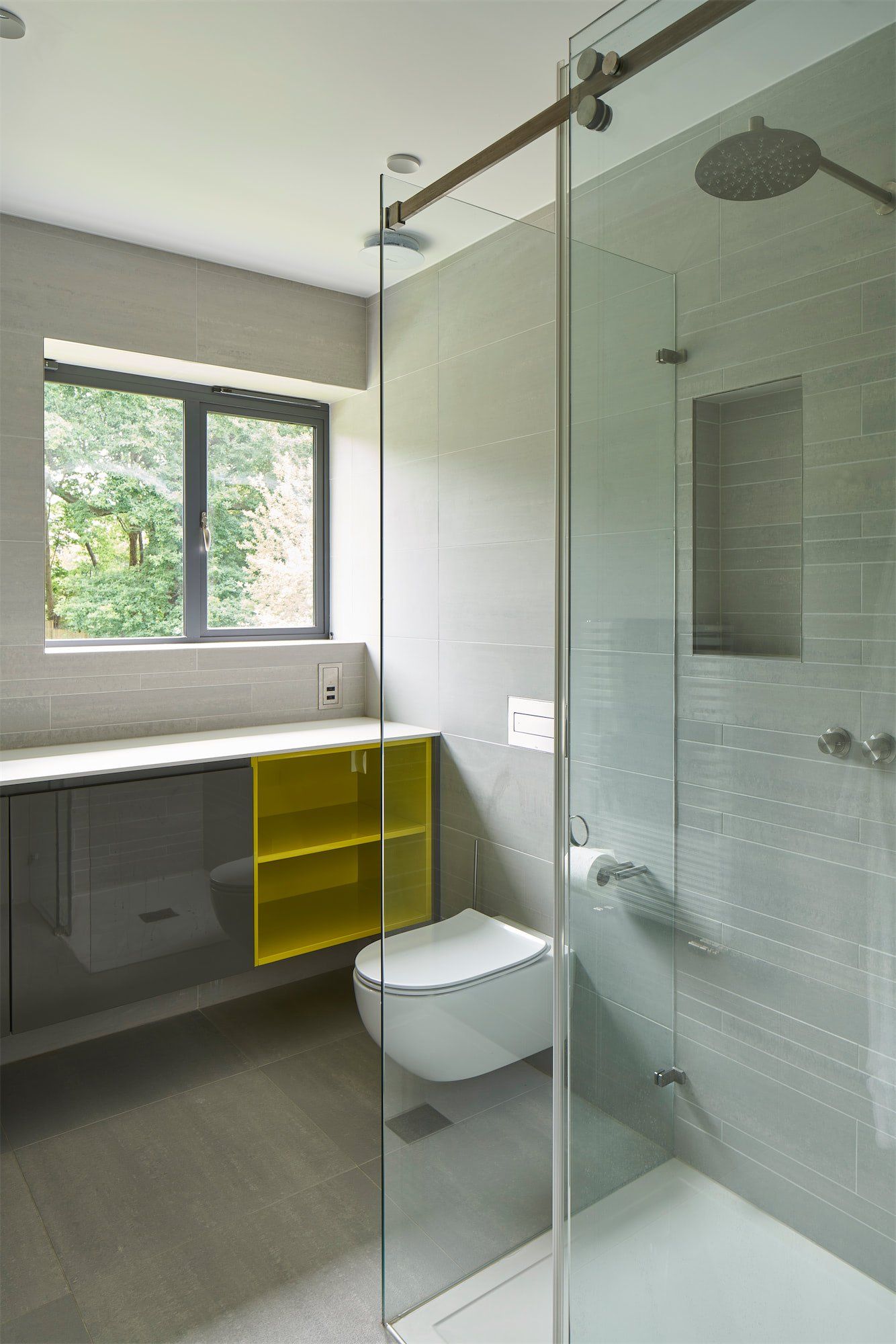 Modern bathroom with glass shower, wall-mounted toilet, and yellow shelving by a window
