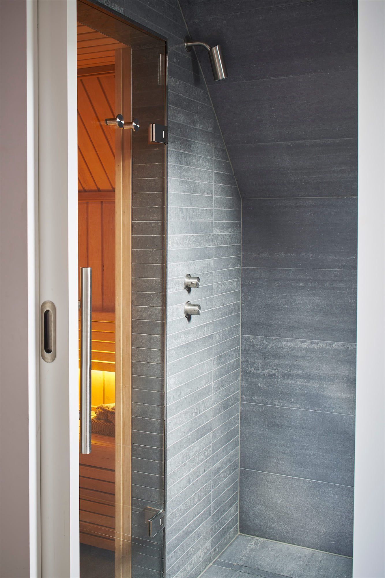 Modern shower stall with gray tile walls and a glass door, next to a warm-lit wooden doorway