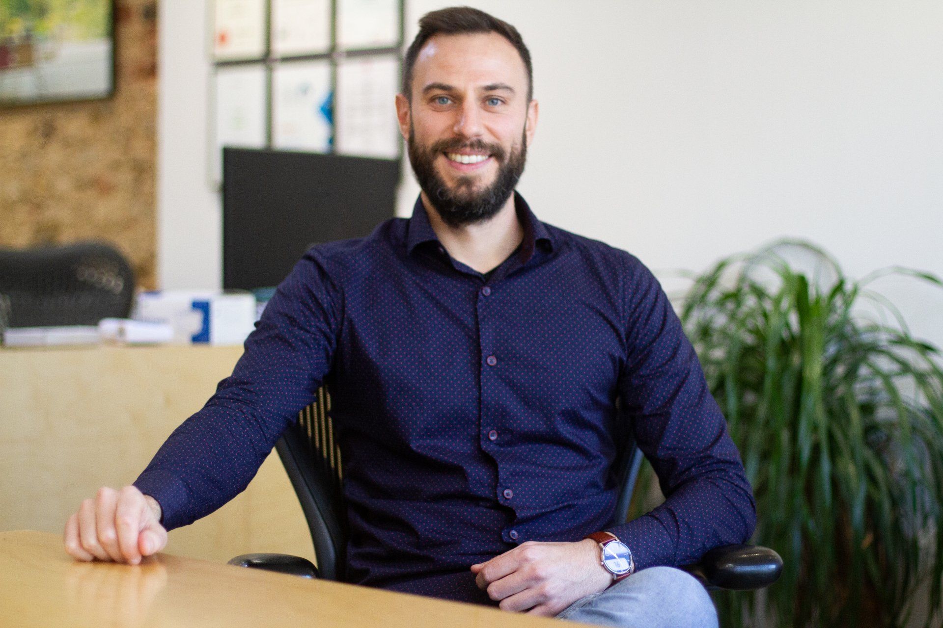 Smiling man in a navy shirt seated at a desk in an office, with plants and a blurred background.