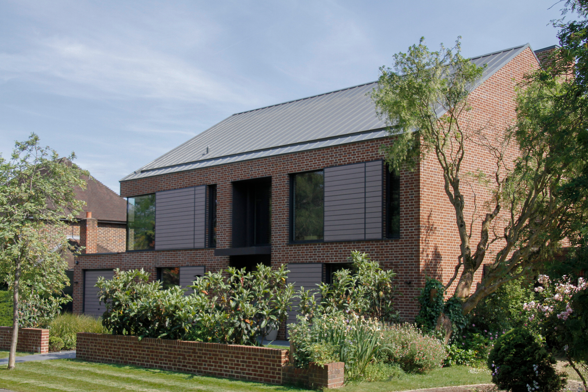 Modern brick townhouse with dark shutters, greenery, and a sloped roof under a clear sky