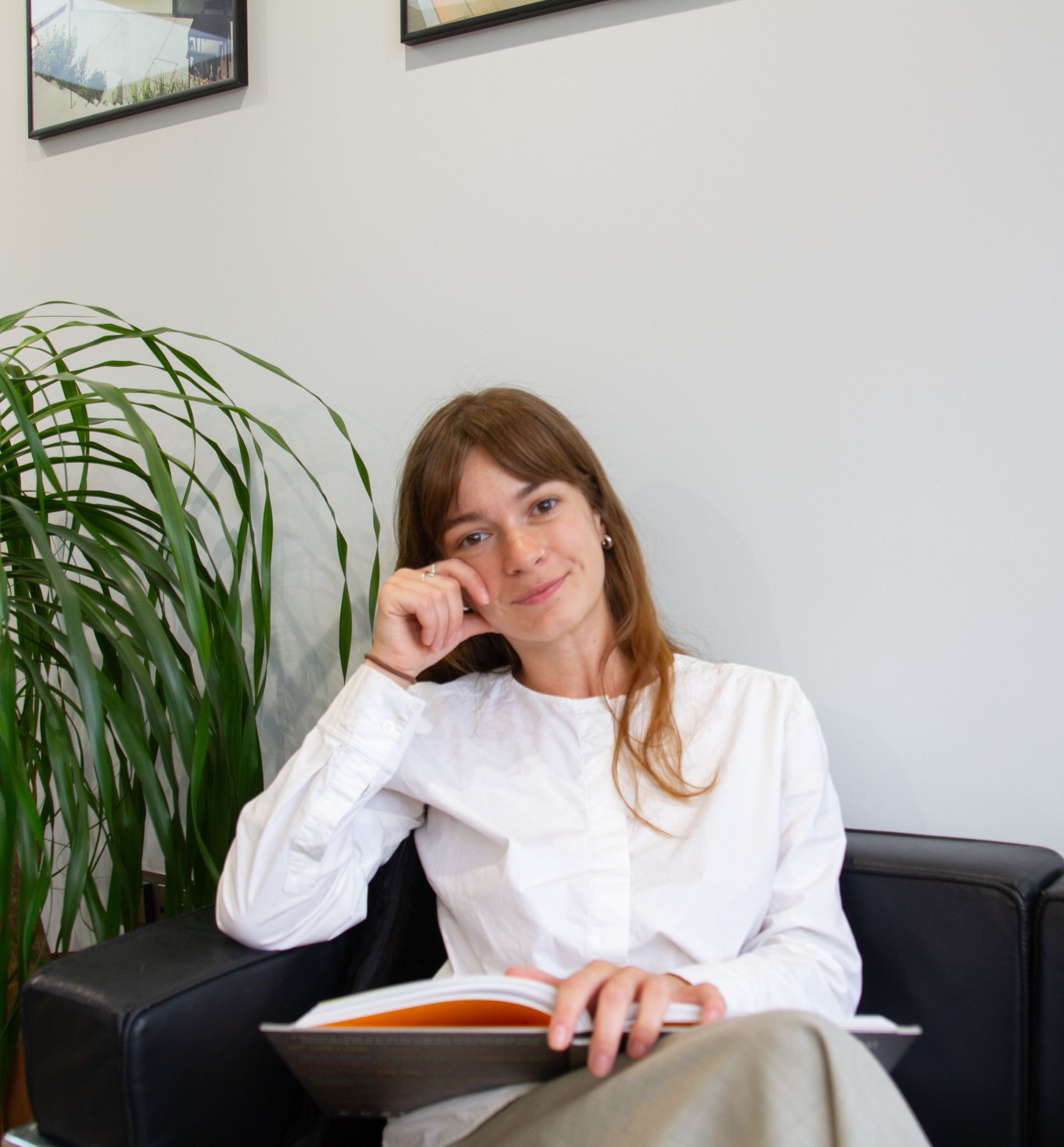 Person in a white shirt sitting on a black couch with a laptop, beside a large potted plant.
