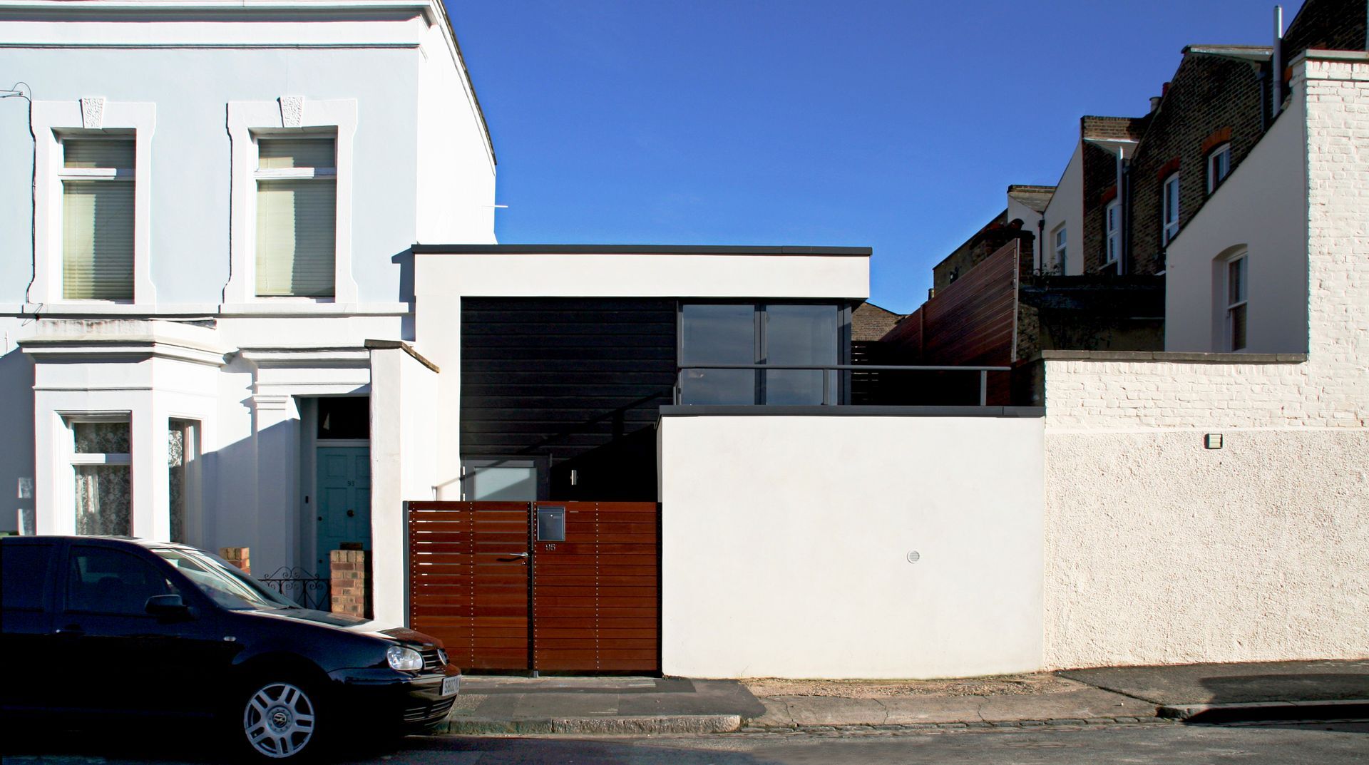 Residential street with white houses, a parked dark car, and a modern white wall in front