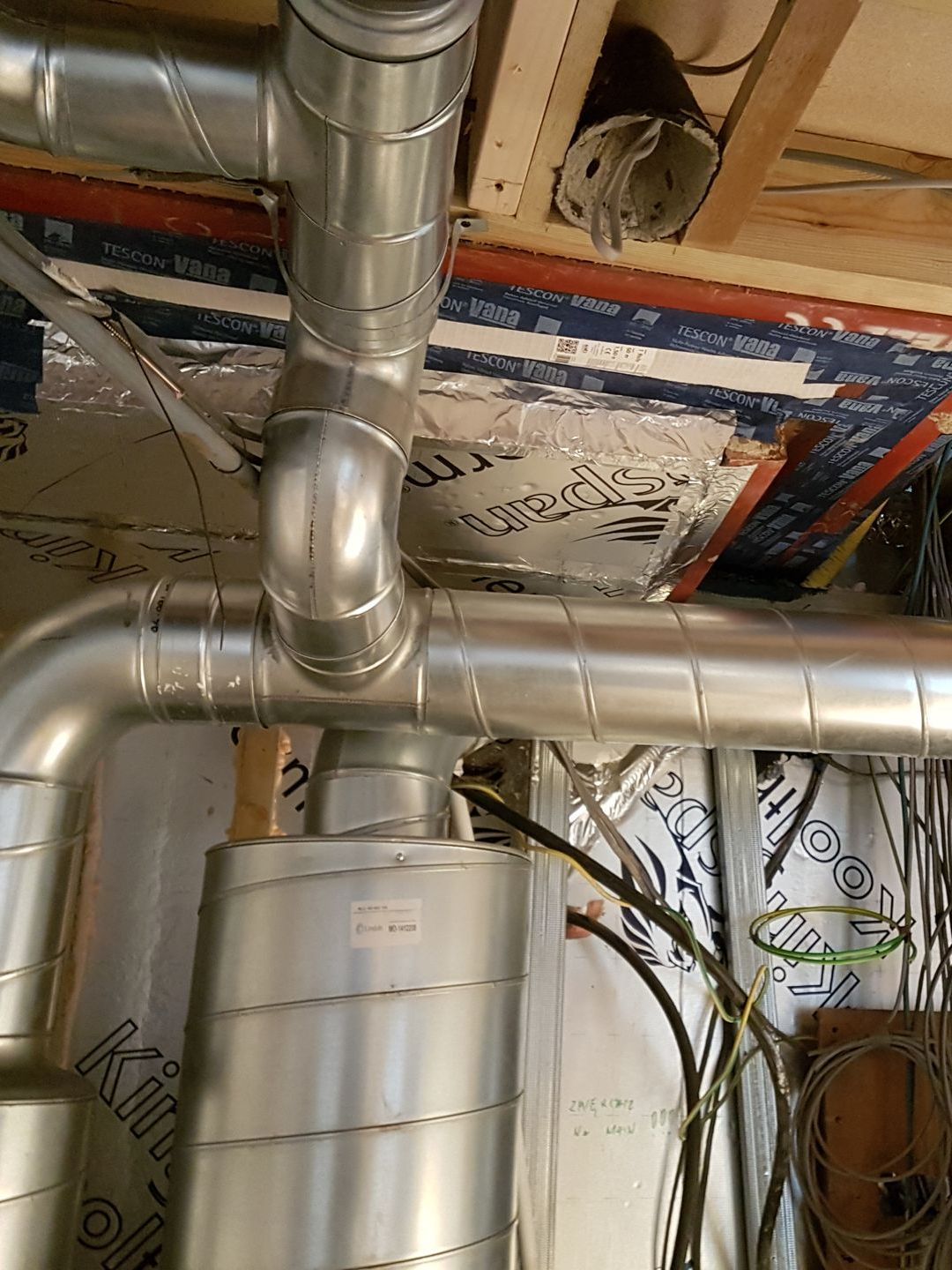 Metal HVAC ducting and a cylindrical vent unit in a basement ceiling with exposed pipes and insulation