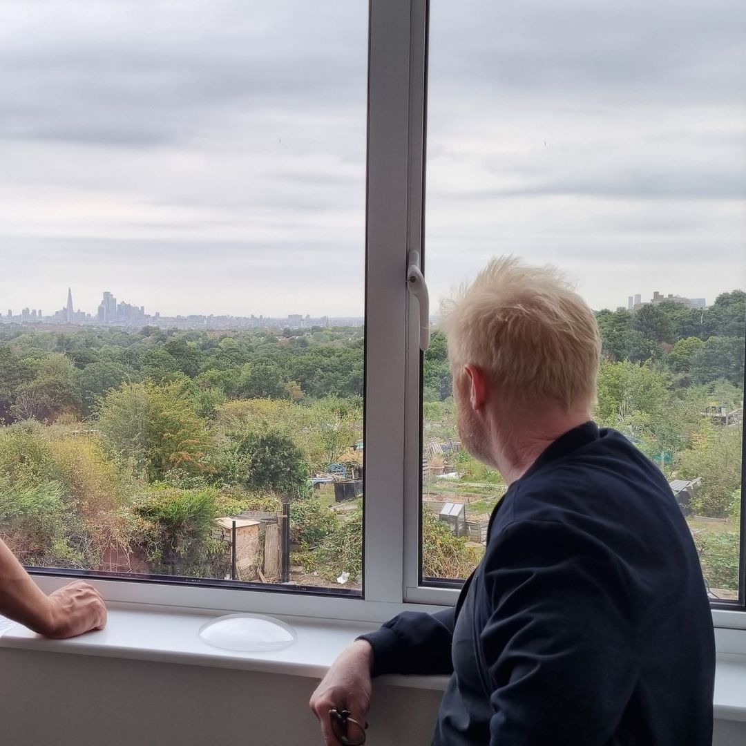Two people look out a window over a green park and distant city skyline under cloudy skies