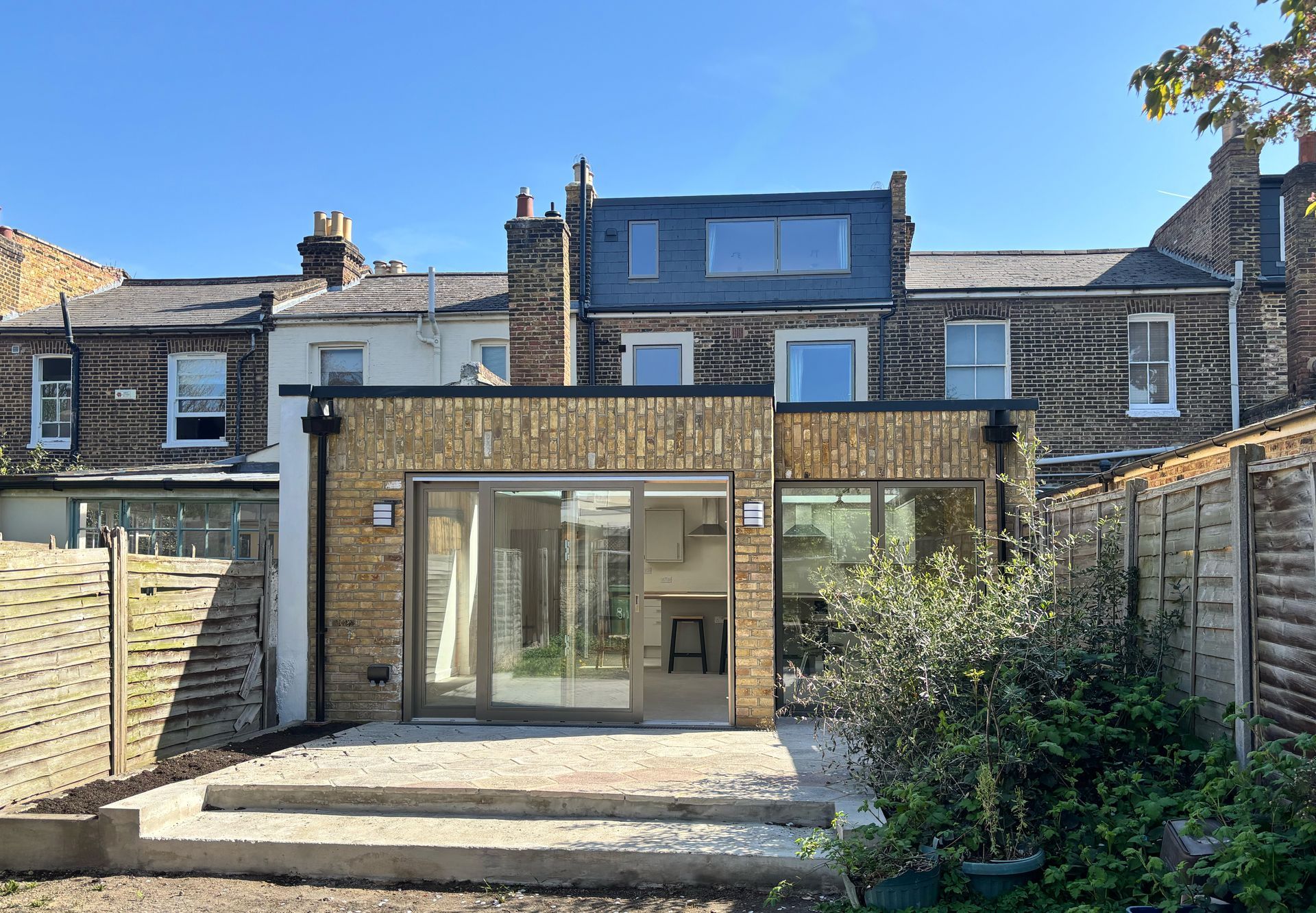 Rear view of a modern brick house with large glass doors, patio, and fenced backyard garden