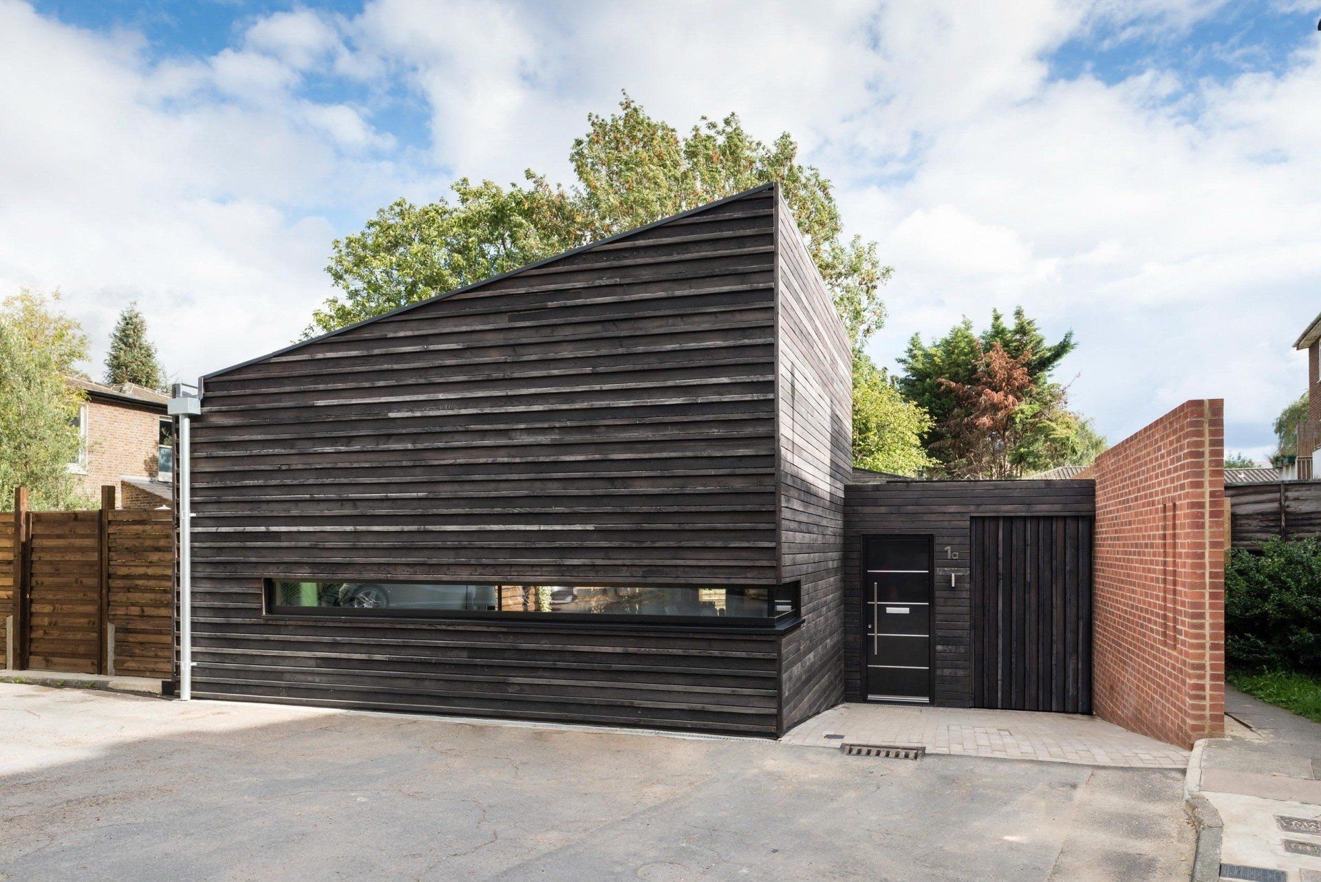 Modern black-clad house with angular roof and brick accents, seen from the street under a partly cloudy sky