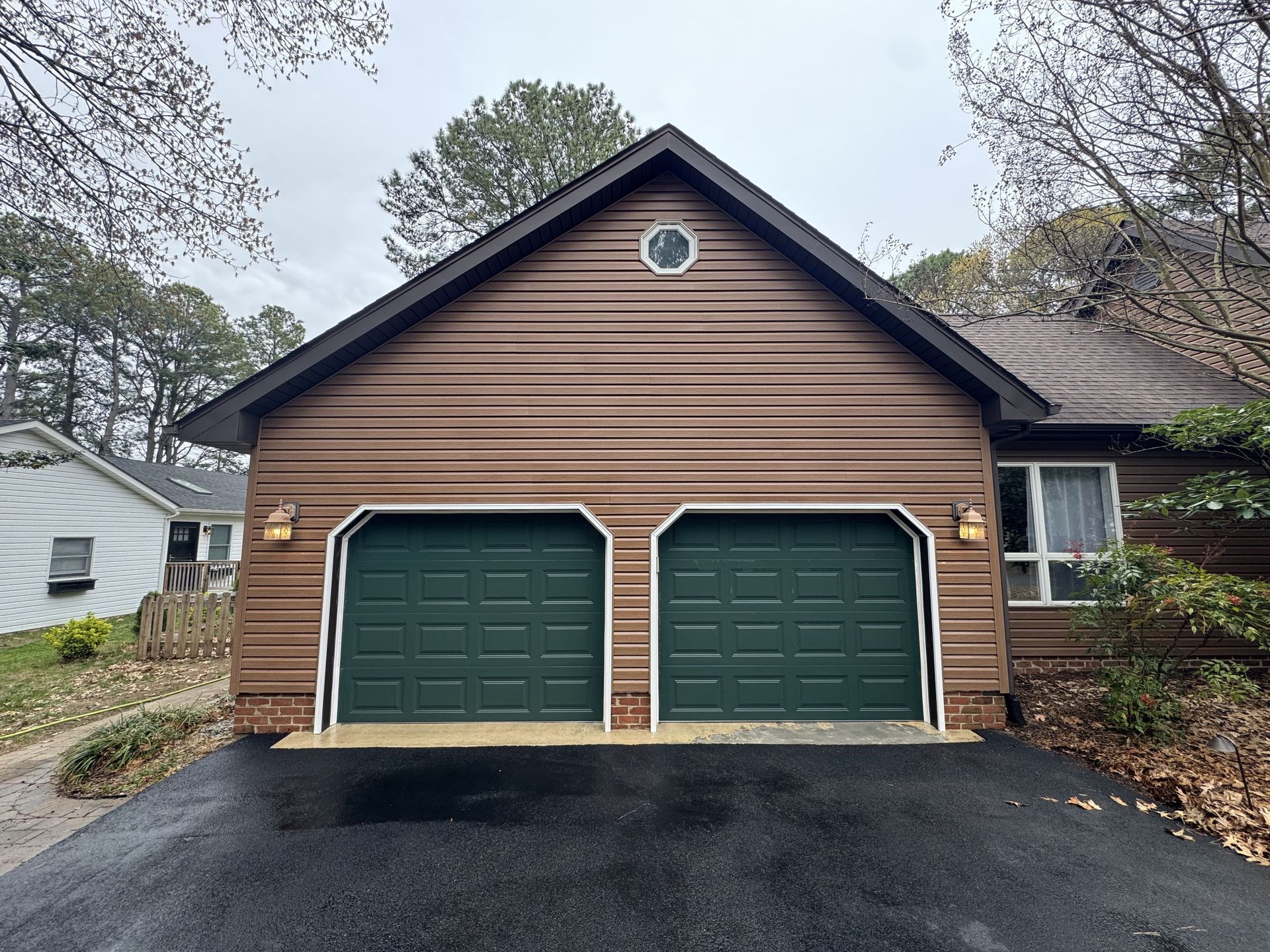 Brown house with two green garage doors. Dark asphalt driveway, cloudy sky.