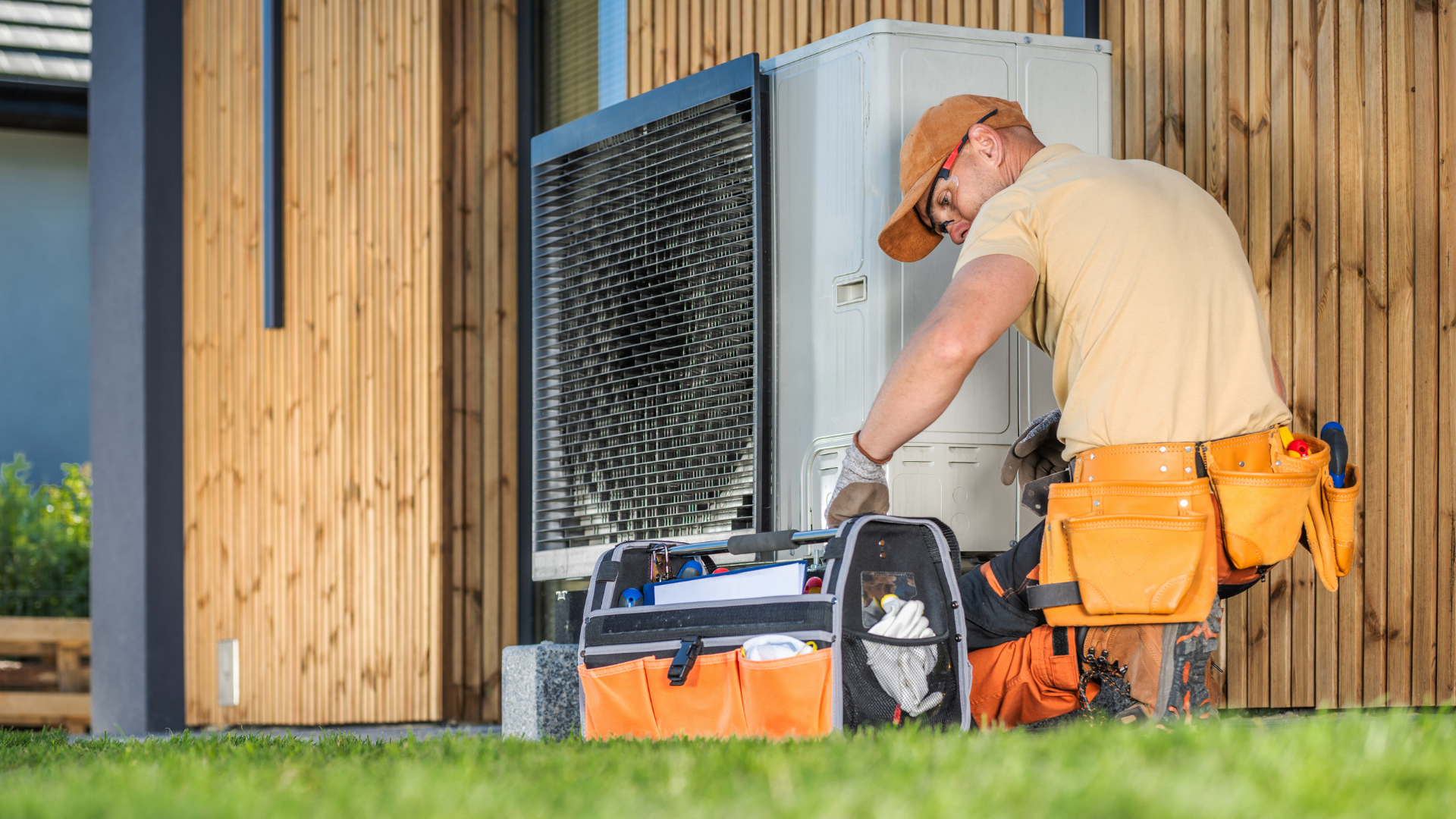 Worker in orange tool belt adjusting an outdoor HVAC unit beside a wooden building