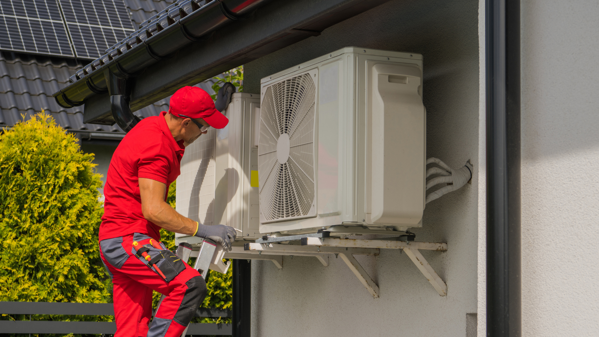 Technician in red uniform servicing an outdoor air conditioning unit on a house wall