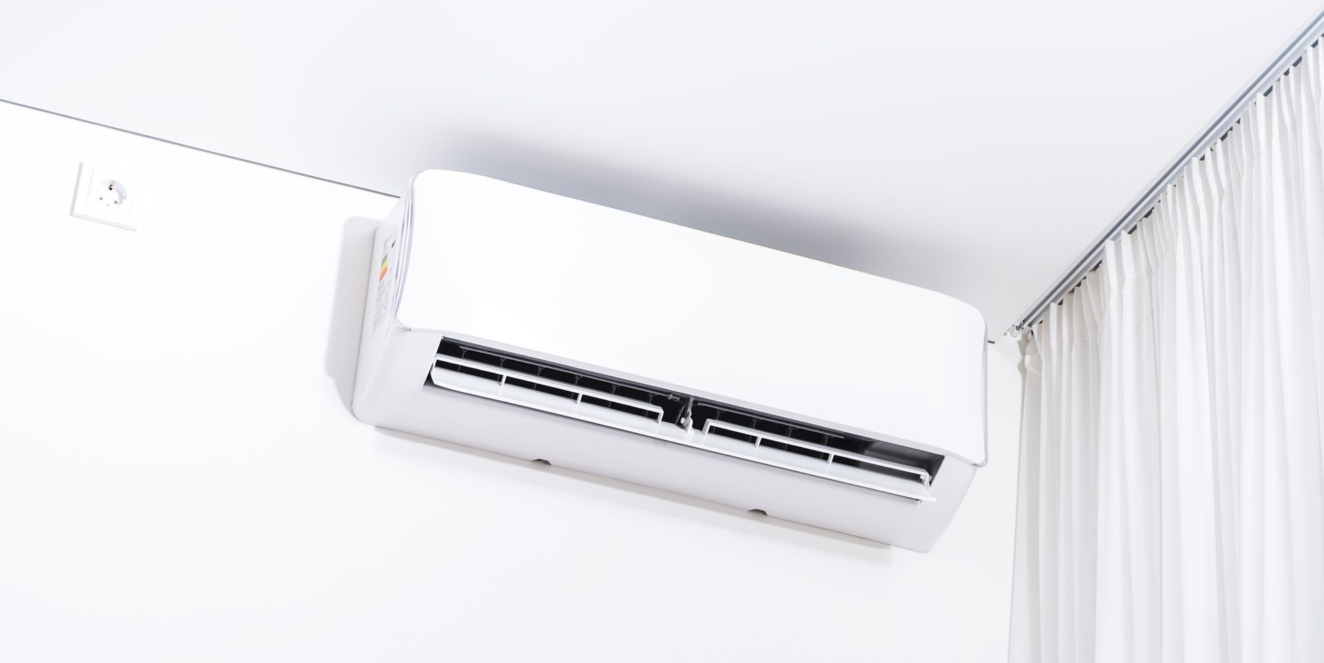 White air conditioning unit on a white wall near white curtains.