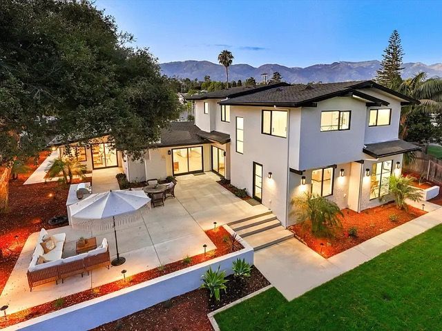 Modern two-story house with lit windows, patio seating, and landscaped yard at dusk