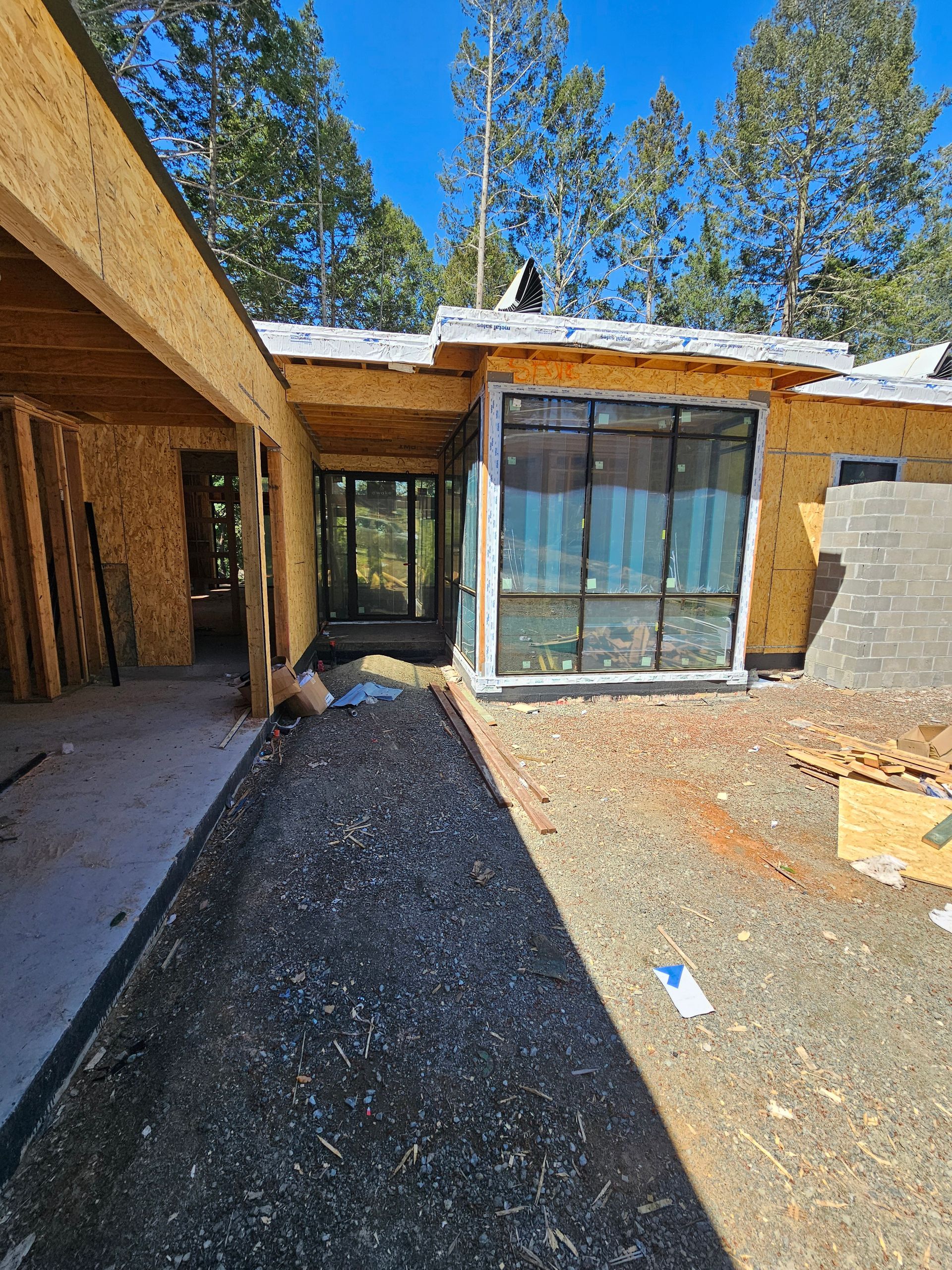 Partially built house exterior with gravel yard, large window, and covered walkway in bright sun