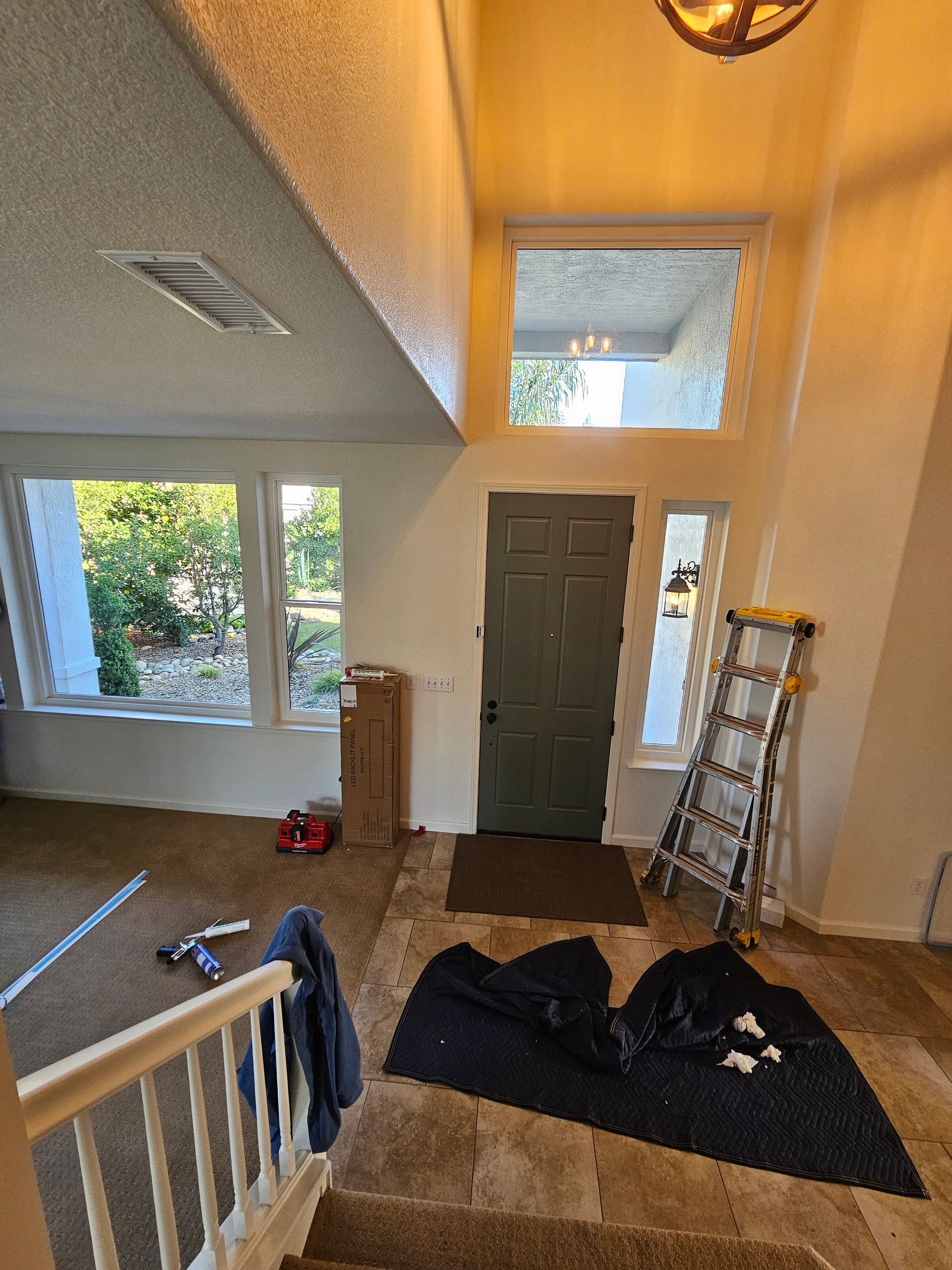 Interior entryway with a green front door, tall window, staircase, coat rack, and ladder on a dark rug.