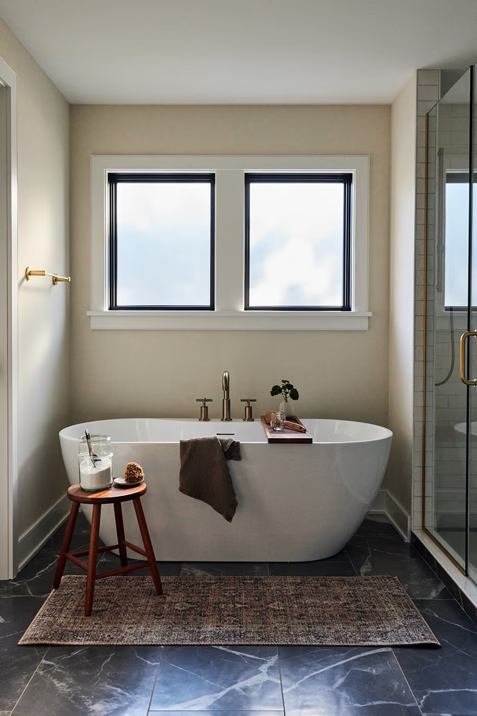 Modern bathroom with freestanding tub, double windows, glass shower, and dark tiled floor