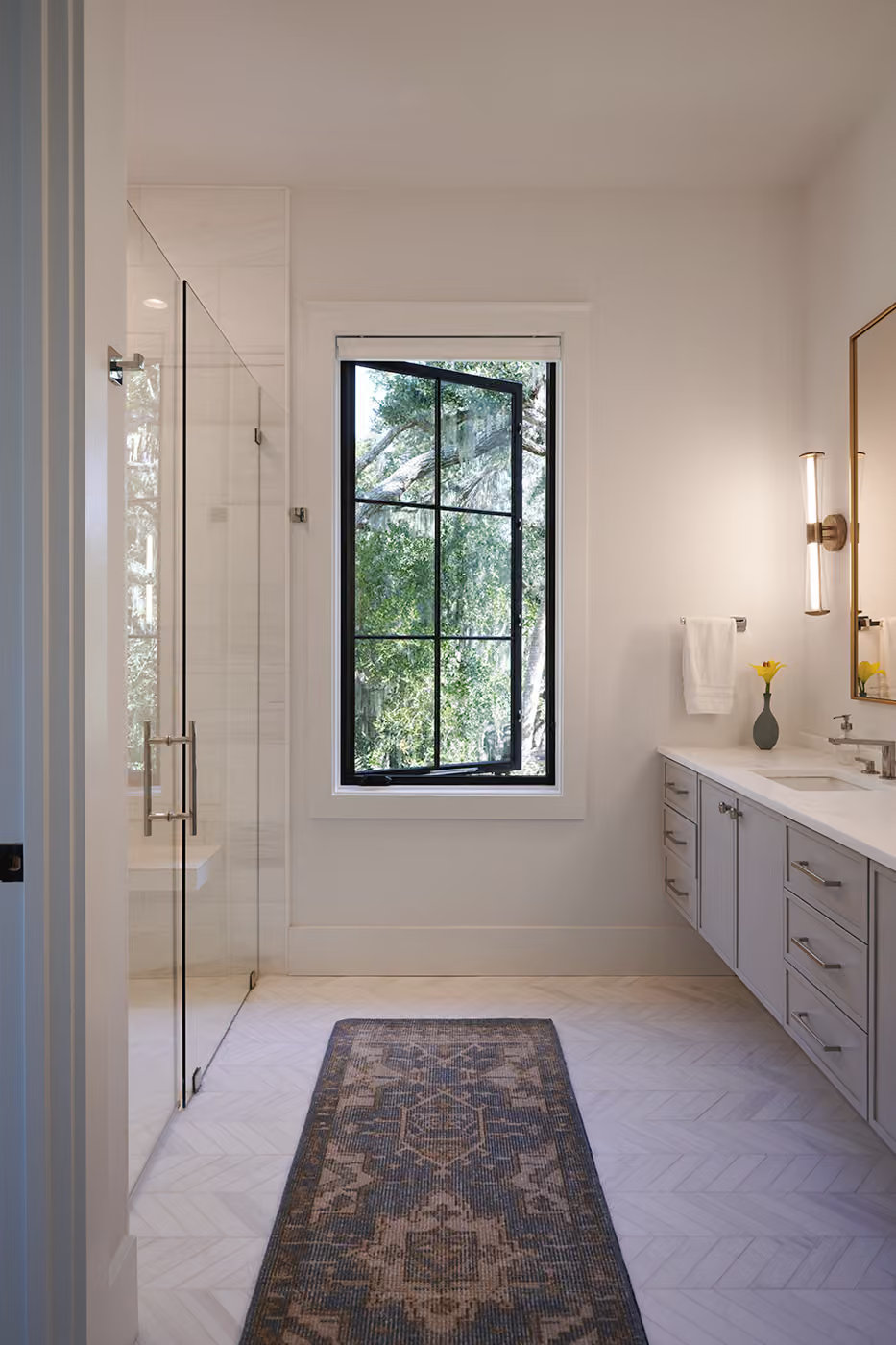 Modern bathroom with glass shower, vanity, window, and patterned runner rug.