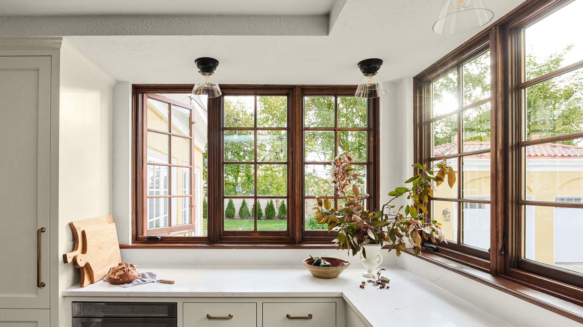 Bright sunroom desk with large windows, white walls, and potted plants overlooking greenery