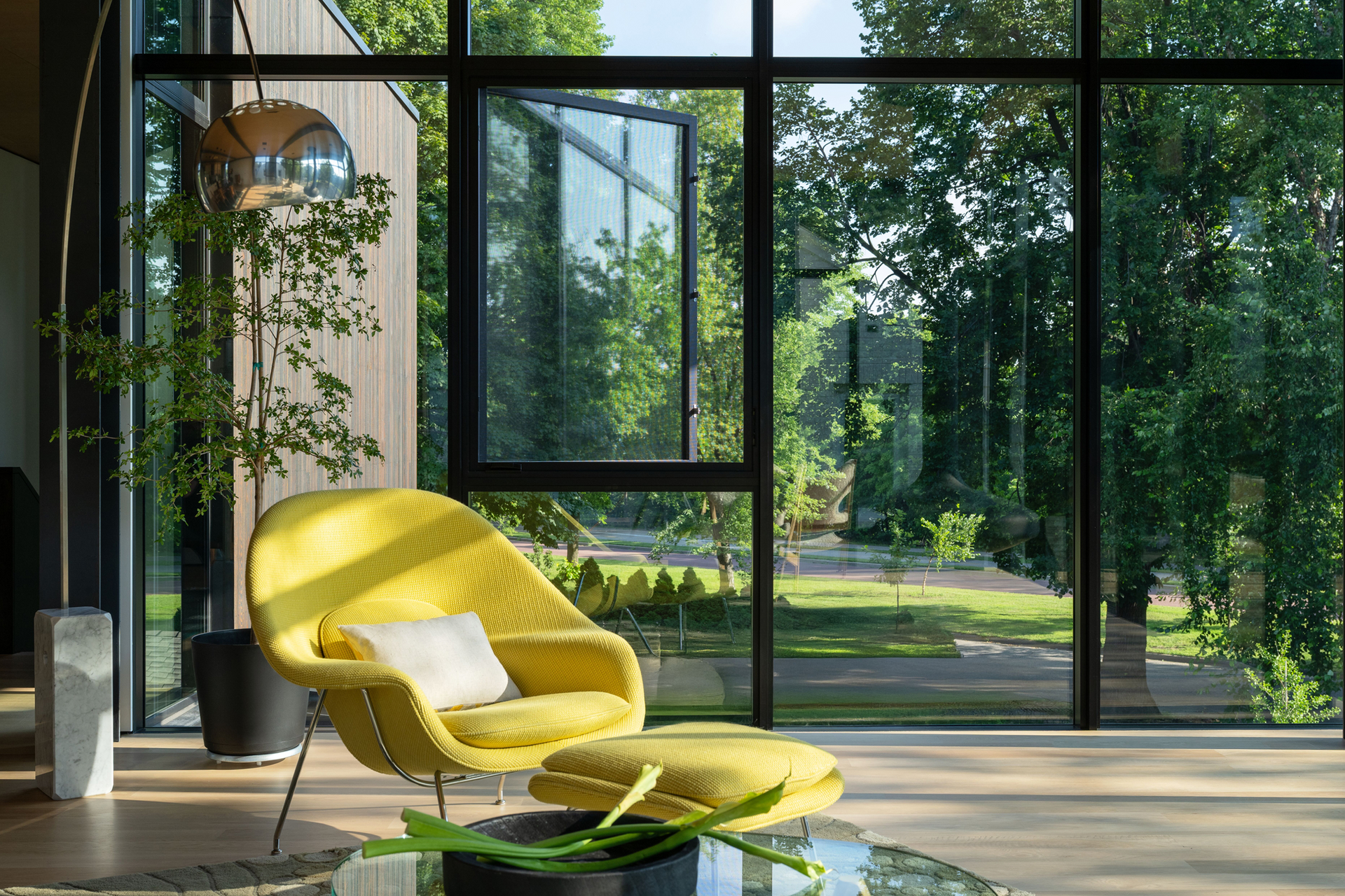 Modern living room with a yellow chair, glass coffee table, and floor-to-ceiling windows overlooking a green garden