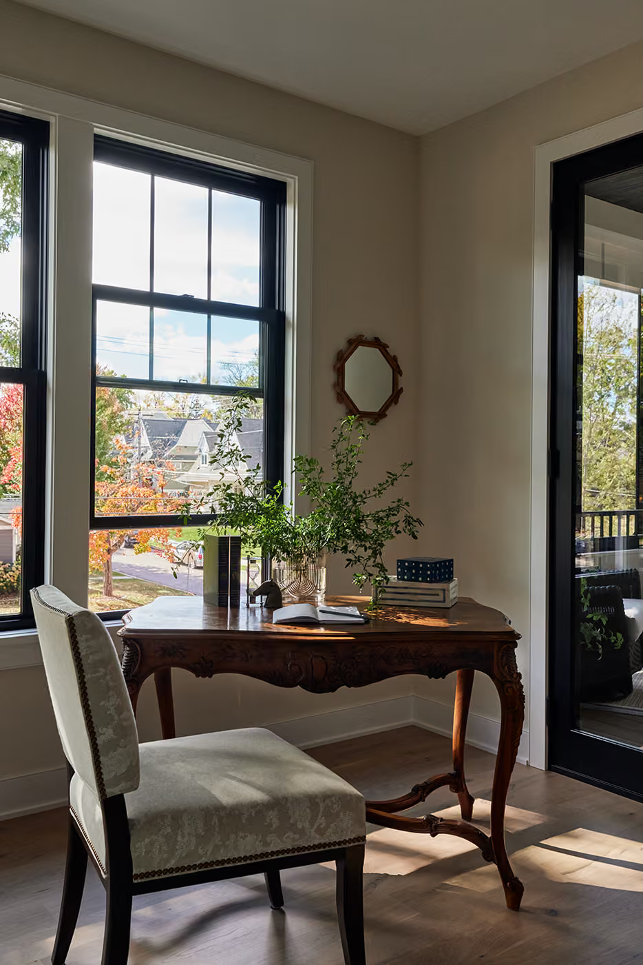 Sunlit room with a wooden desk, chair, plant, and tall windows beside a glass door.