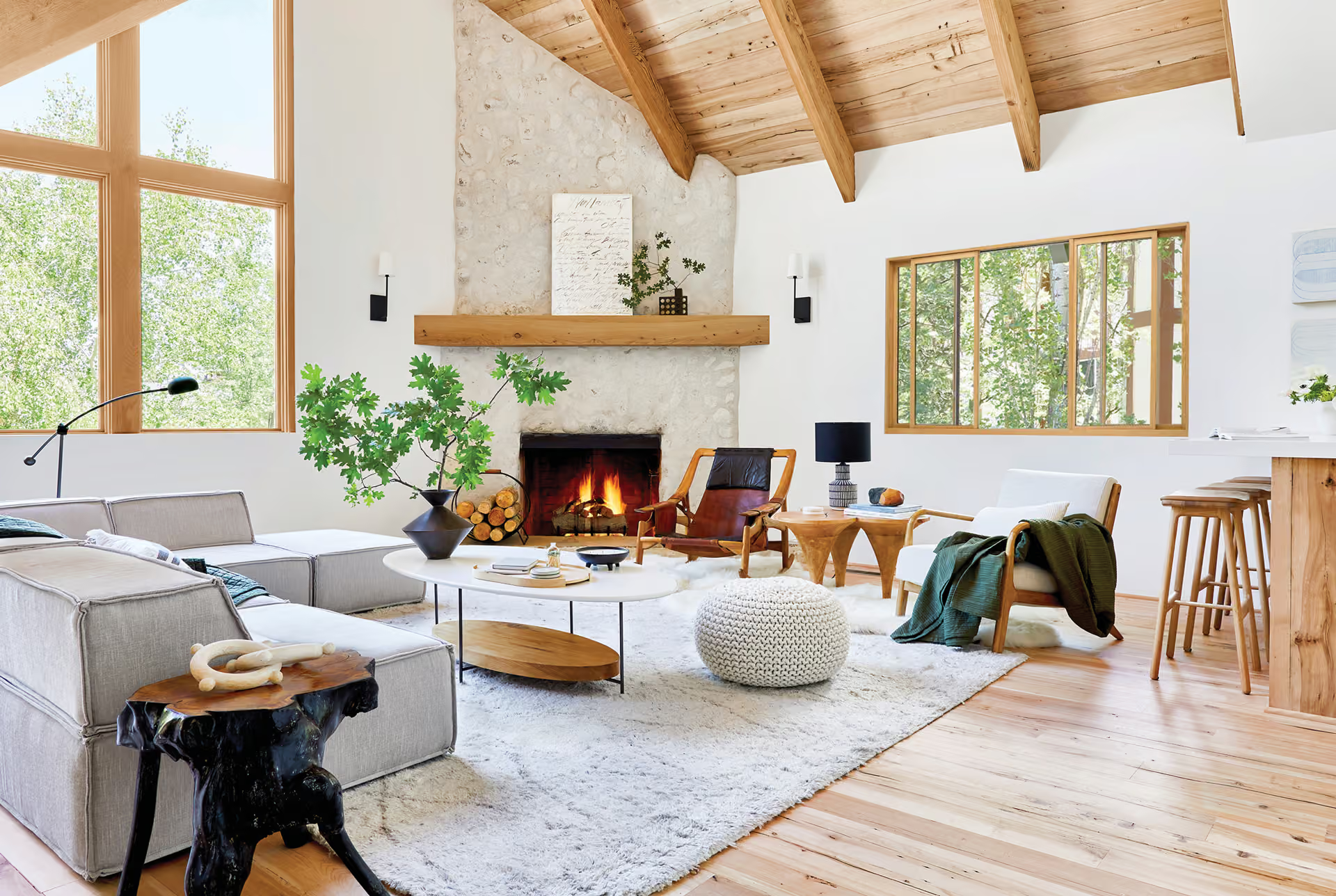 Bright living room with vaulted wood ceiling, fireplace, gray sofa, and large windows flooding in sunlight