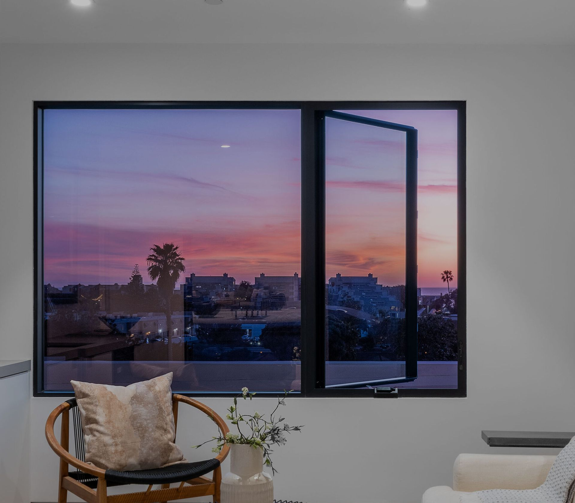 Open window at dusk with pink sky, city silhouette, and a cozy chair by the window