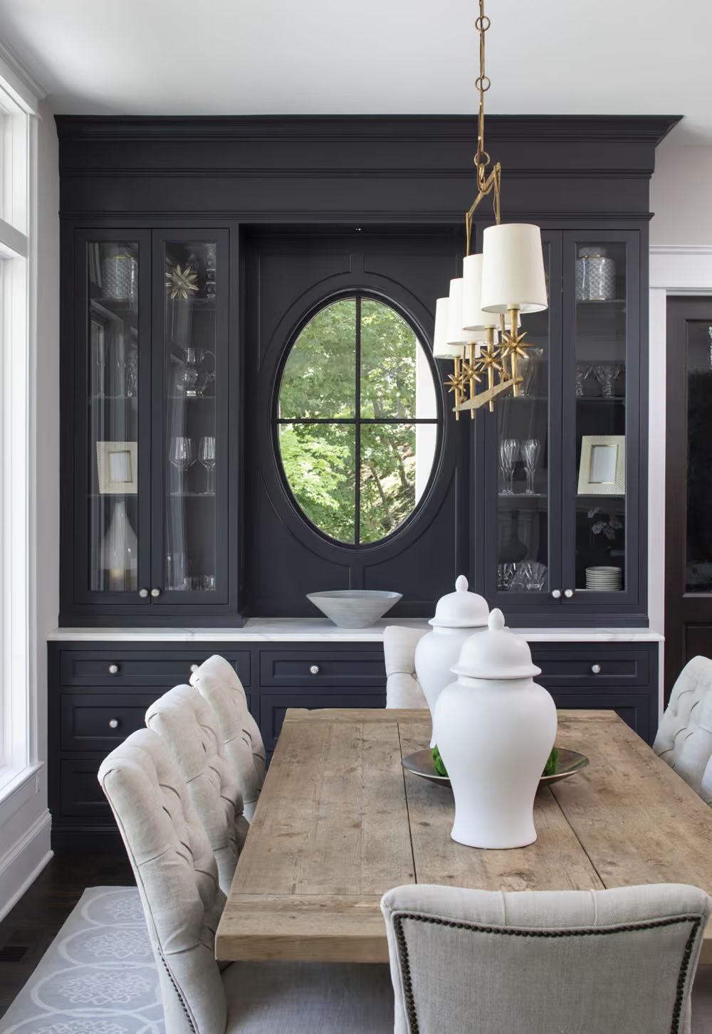 Elegant dining room with black built-ins, round mirror, rustic wooden table, and white chairs