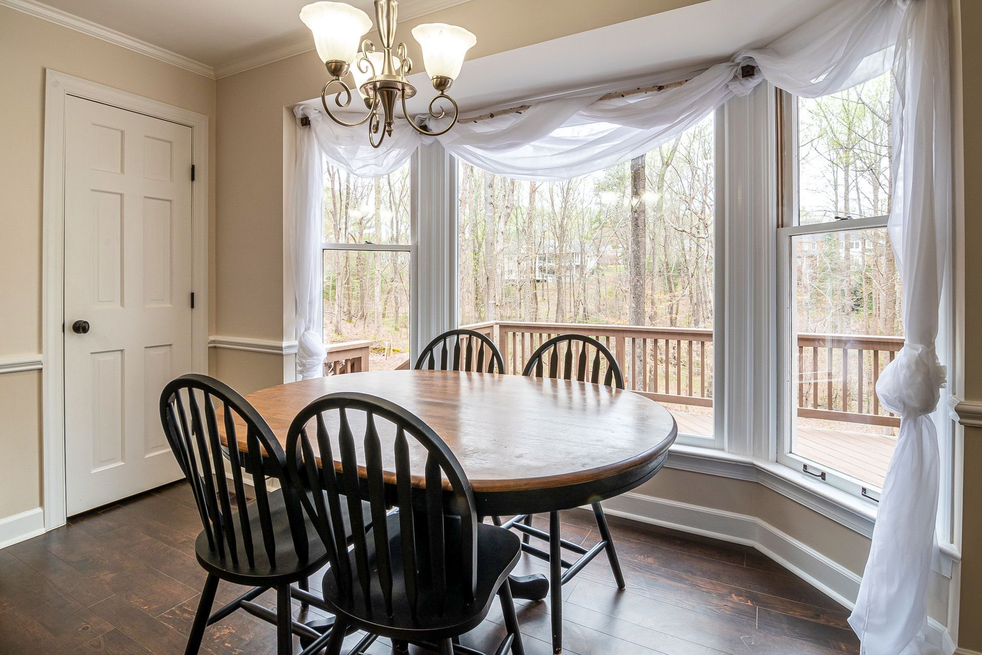 Dining nook with round wooden table, black chairs, chandelier, and large bay windows with sheer curtains.