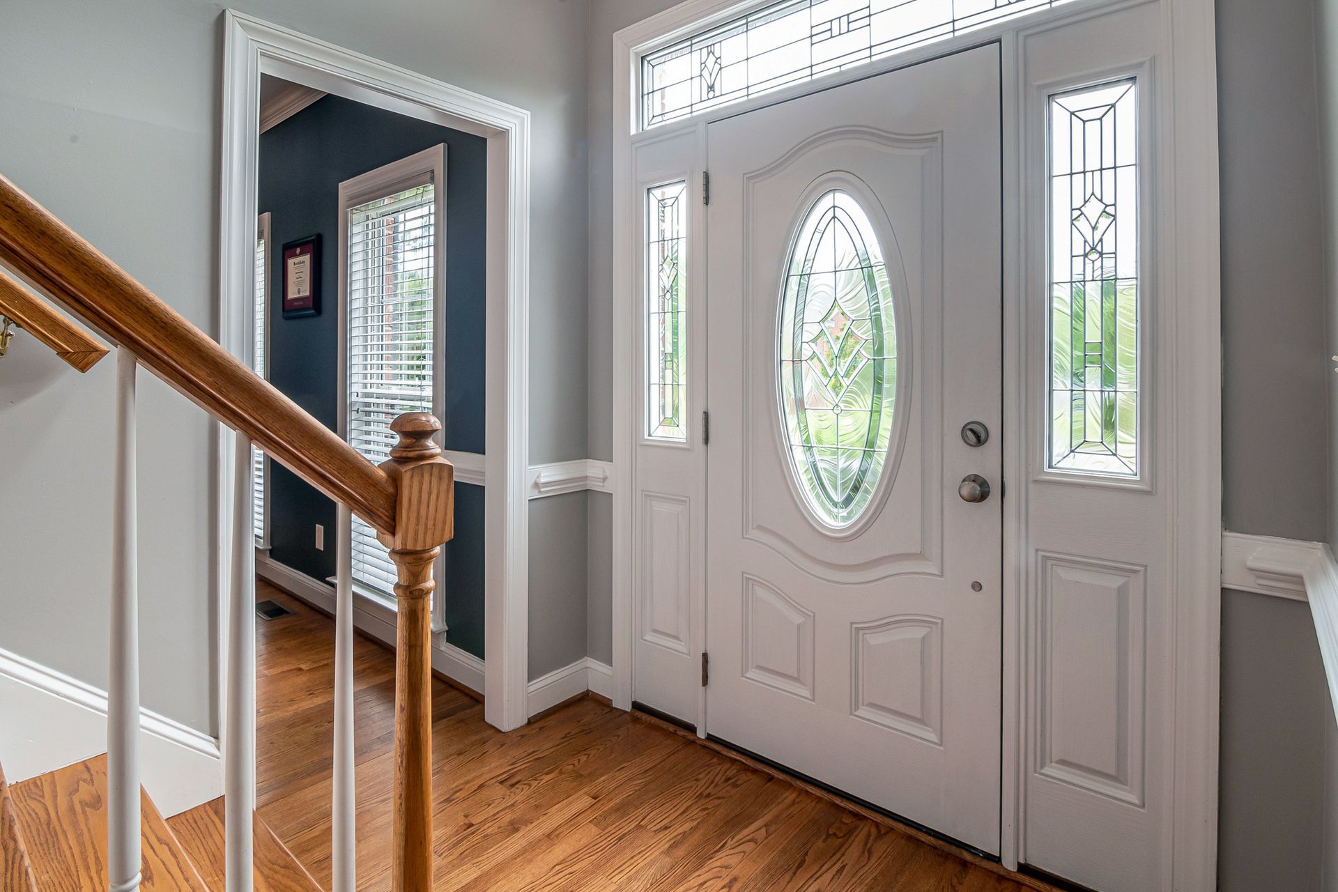 Front entryway with white front door, sidelights, gray walls, and hardwood floor beside a staircase.