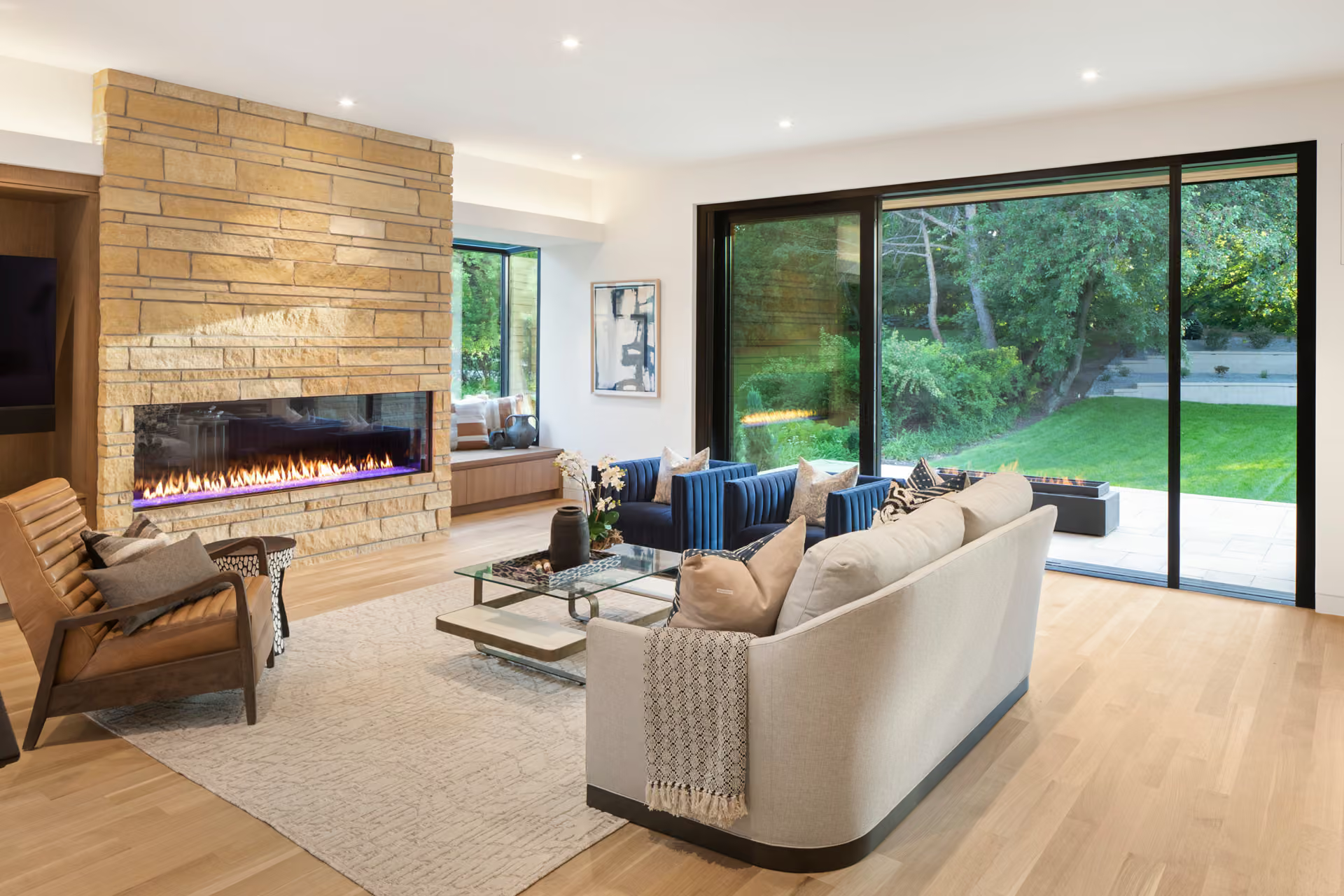 Modern living room with stone fireplace, neutral sofa, and large sliding glass doors to a green backyard