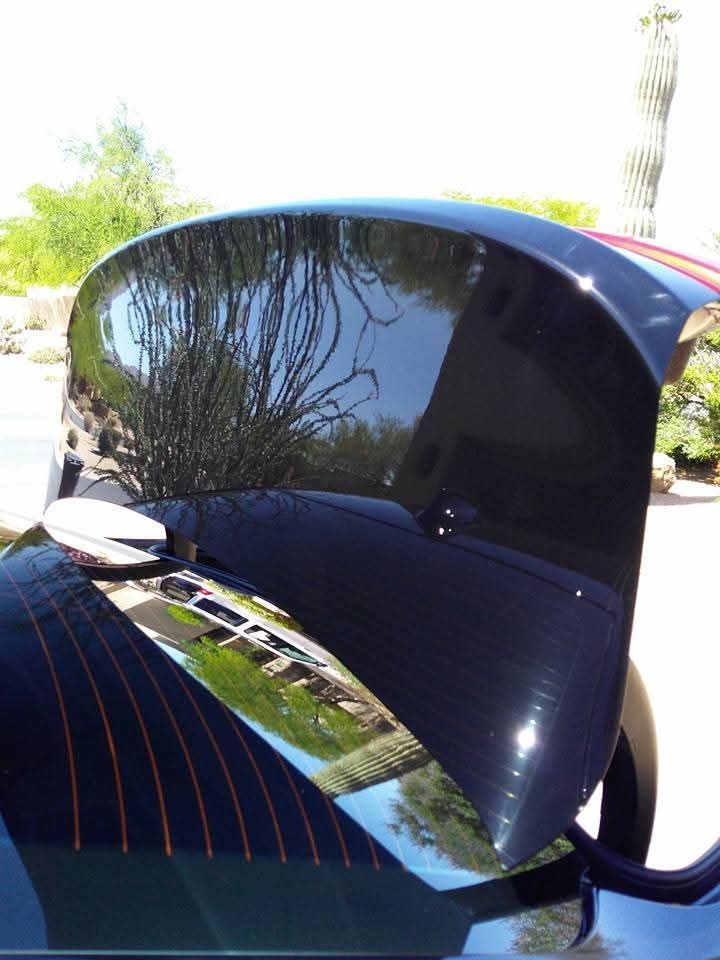 An open, glossy black car trunk lid reflecting the surrounding trees and buildings outdoors.