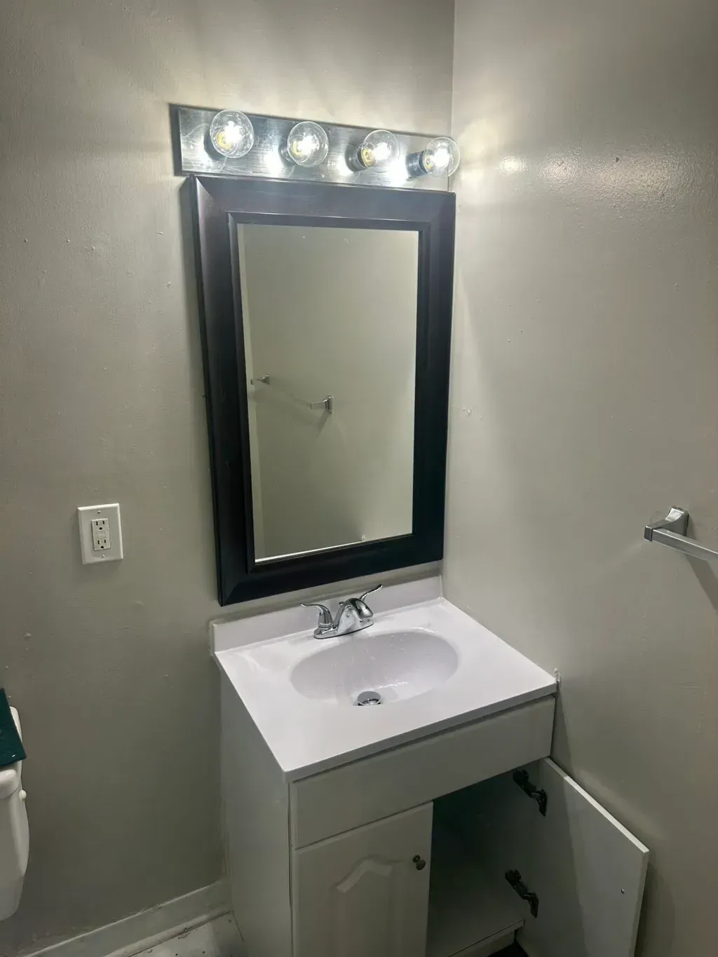 A bathroom vanity with a white cabinet, white countertop and sink, a black-framed mirror, and a four-bulb light fixture.