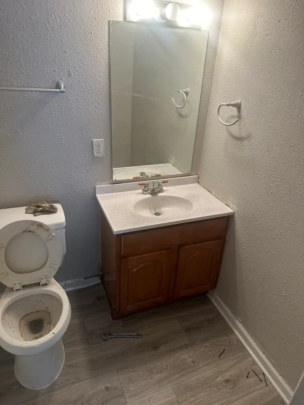A small, simple bathroom with a white toilet, wood vanity, light-colored stone countertop, and mirror on textured walls.