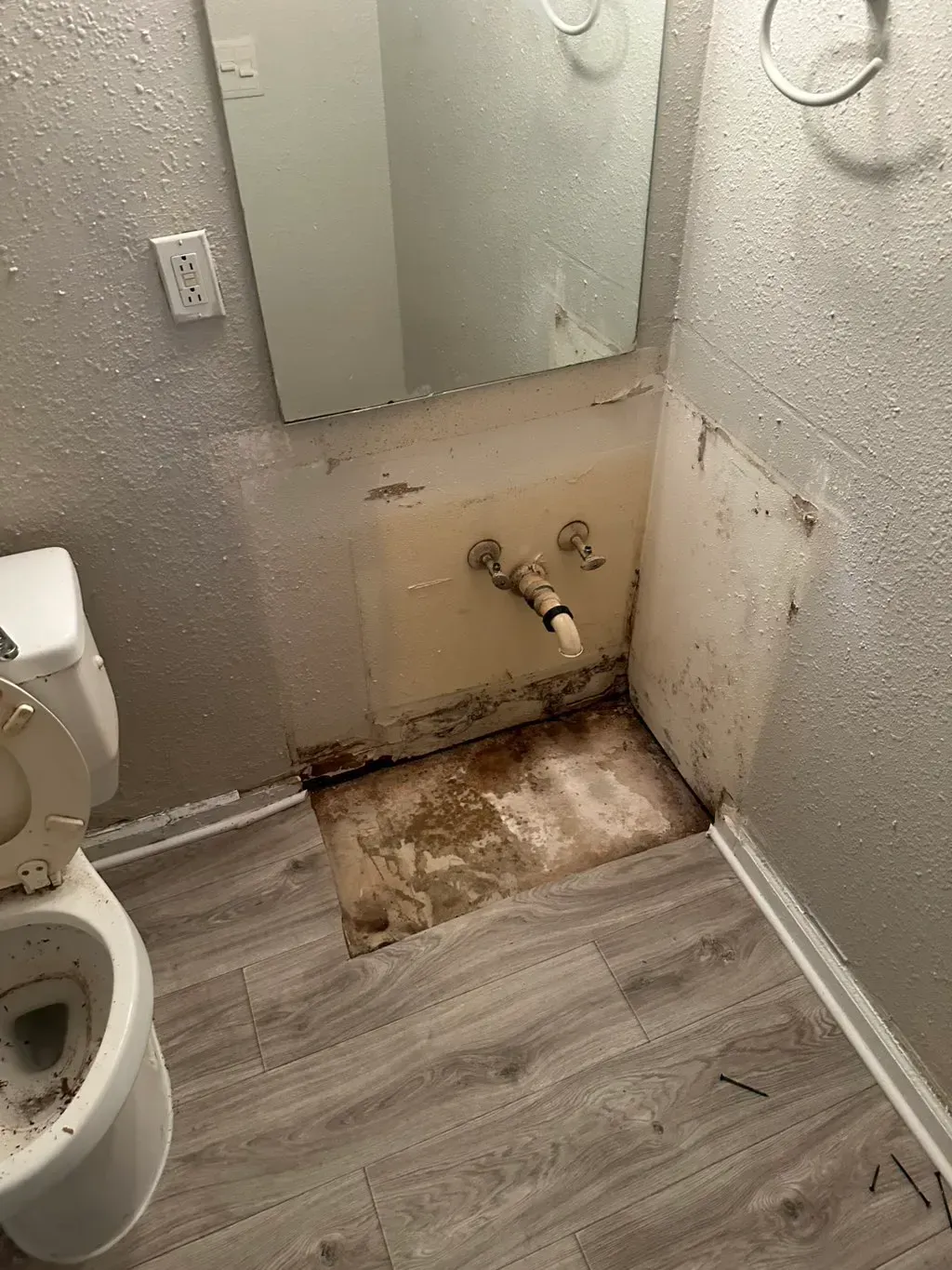 A bathroom sink area under construction with exposed plumbing, damaged drywall, and a removed vanity, next to a toilet.