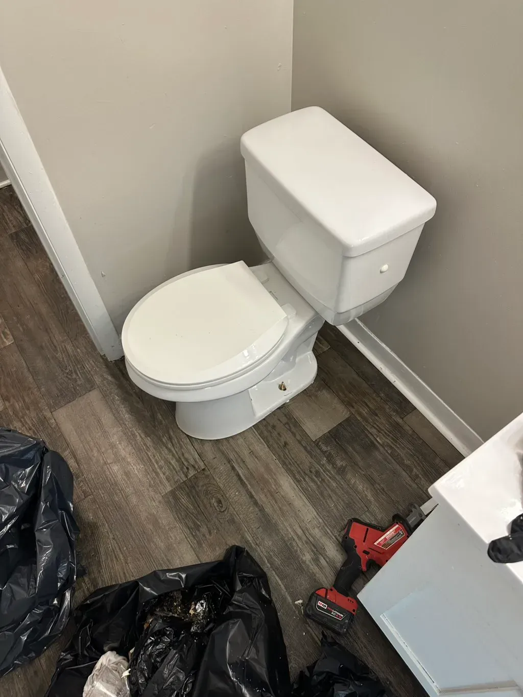 A white toilet in a corner with wood-look flooring and construction tools nearby.