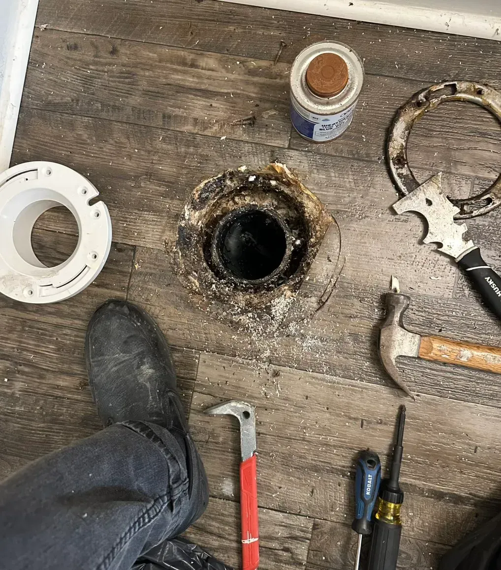 Tools, a new flange, and a damaged toilet drain pipe on a wood-look floor during a bathroom repair.