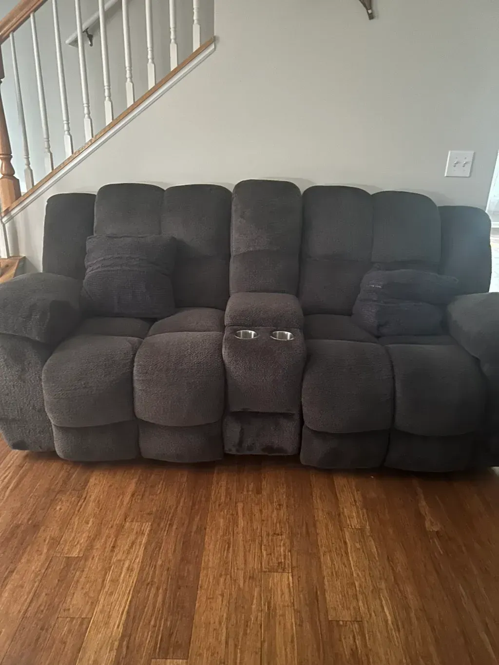 A dark gray, cushioned loveseat with a center console and cup holders, positioned on a light-colored hardwood floor.