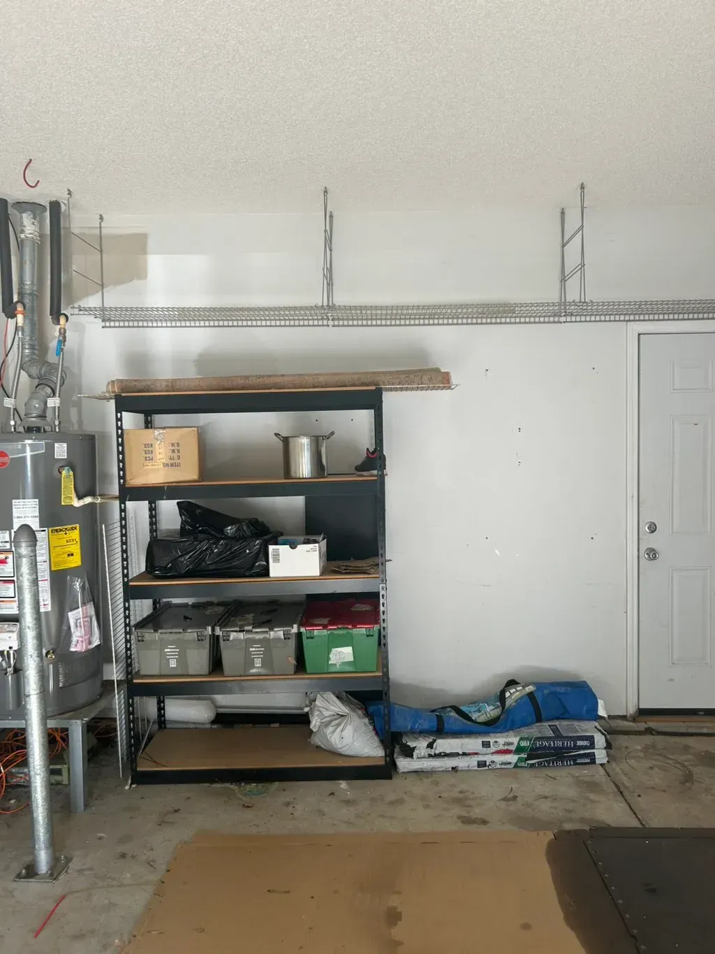 A garage storage area featuring a tall black shelving unit, overhead wire racks, a water heater, and a closed door.