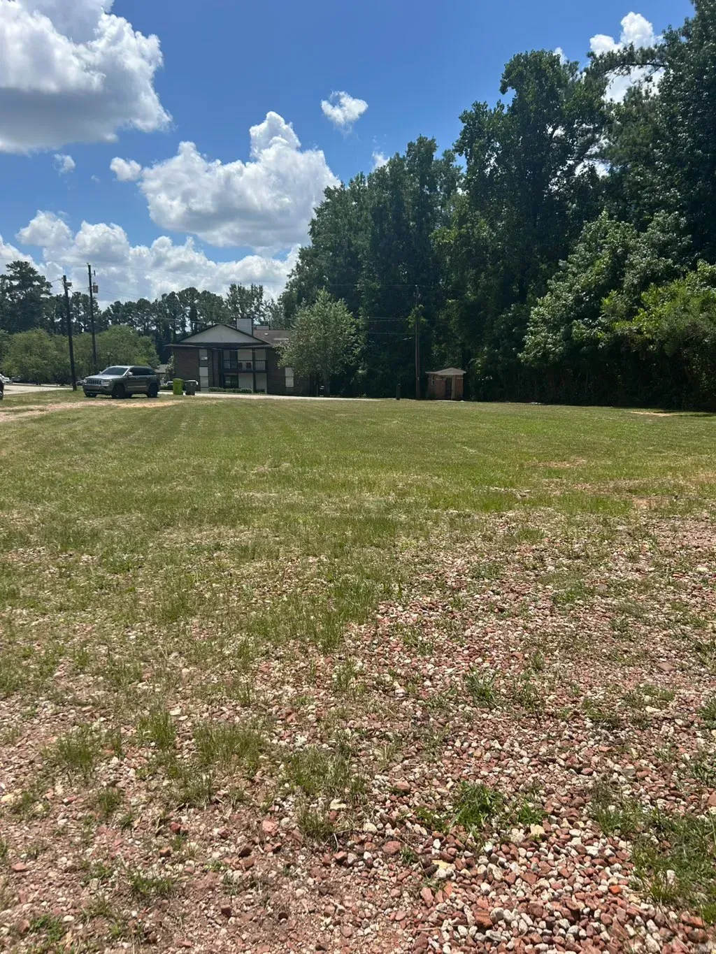 A large, open grassy lot under a blue sky with scattered clouds, bordered by trees and a distant building.