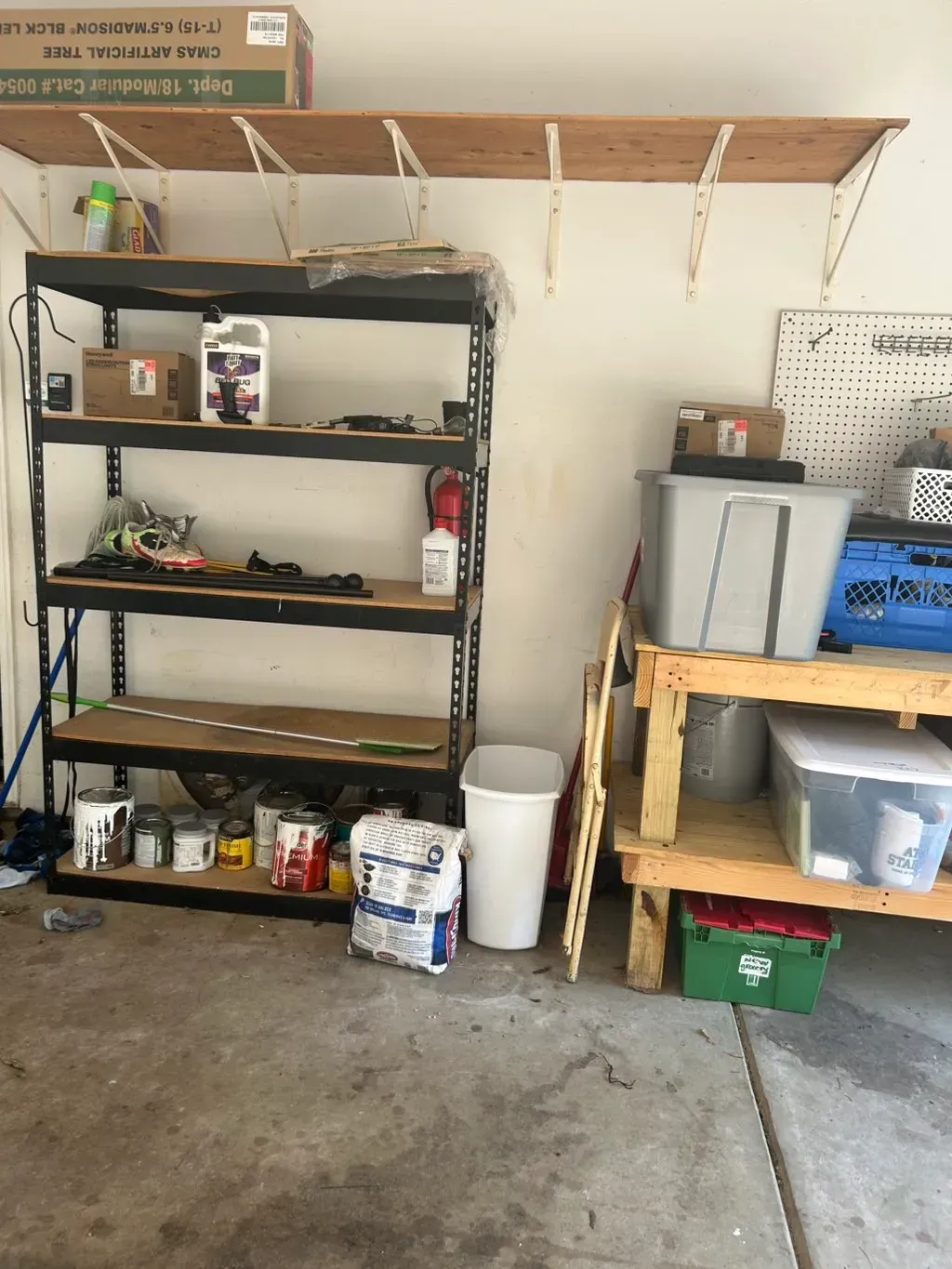 A garage storage area with a black metal shelving unit, a wooden workbench, a wall shelf, and various bins and supplies.