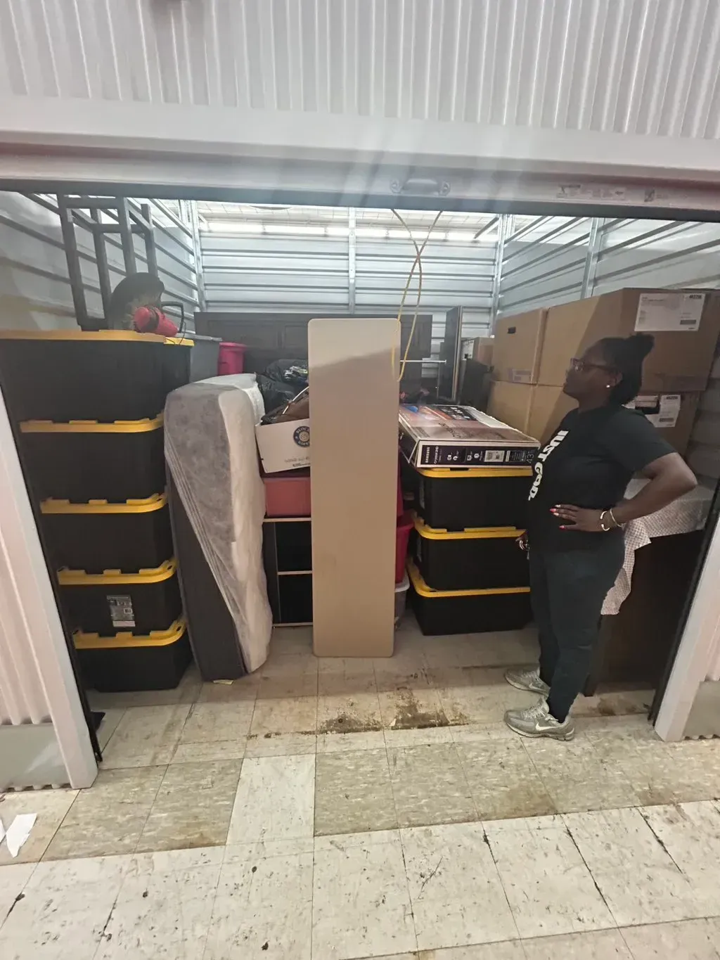A person stands inside a storage unit filled with stacked black plastic storage bins, furniture, and cardboard boxes.