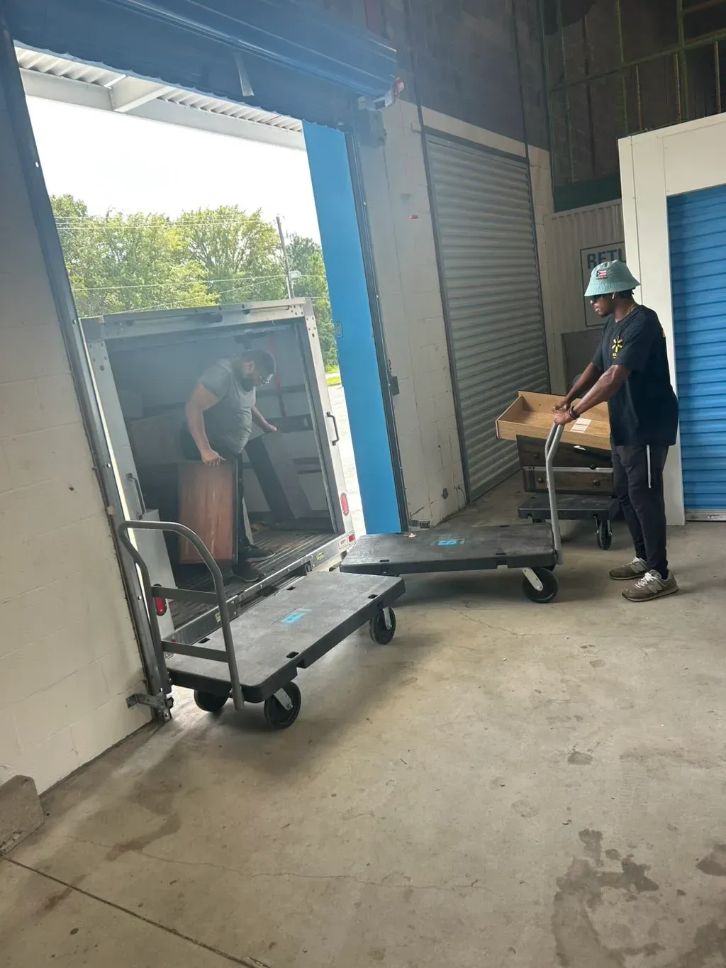 Two people use flatbed carts to unload items from a parked truck into a storage facility.