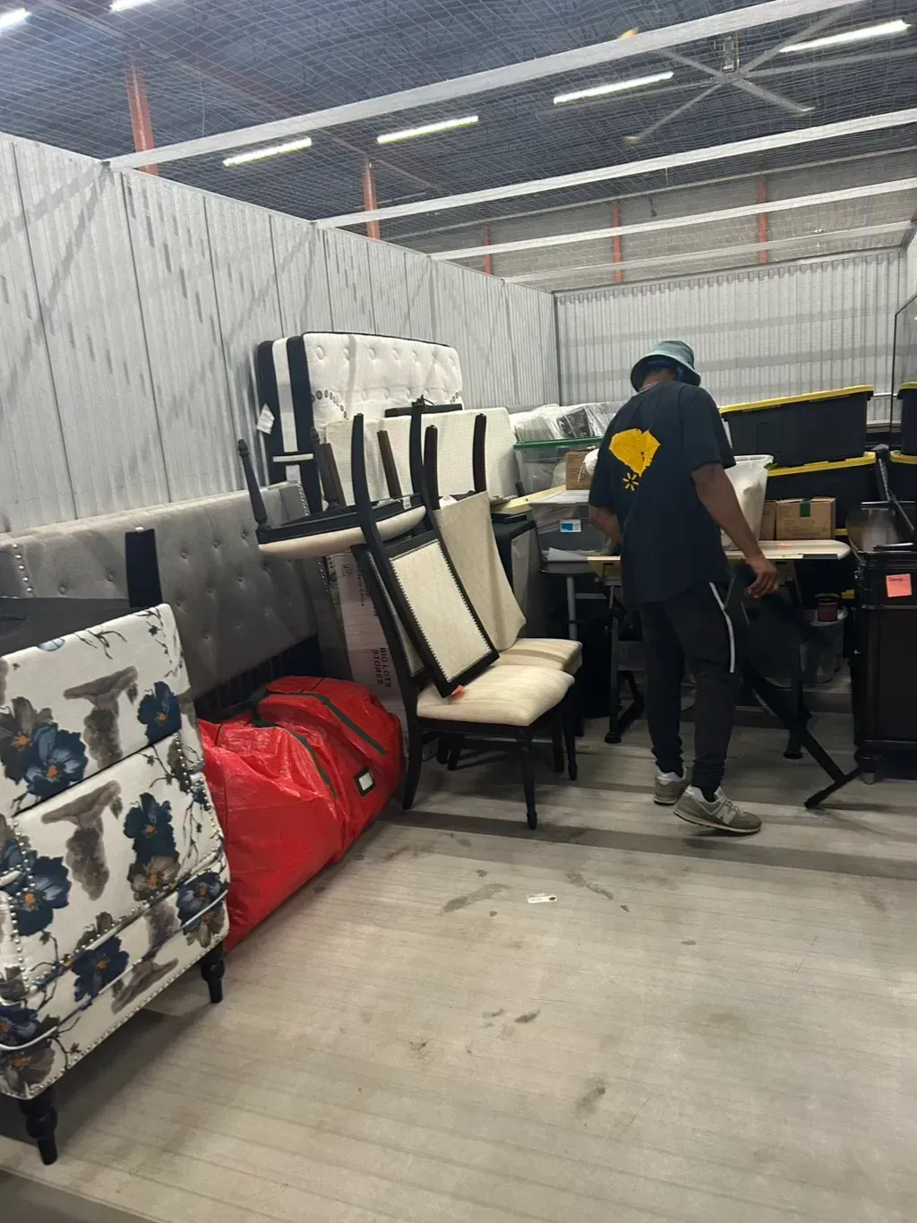 A person in a dark uniform and bucket hat works inside a storage unit filled with packed furniture and chairs.