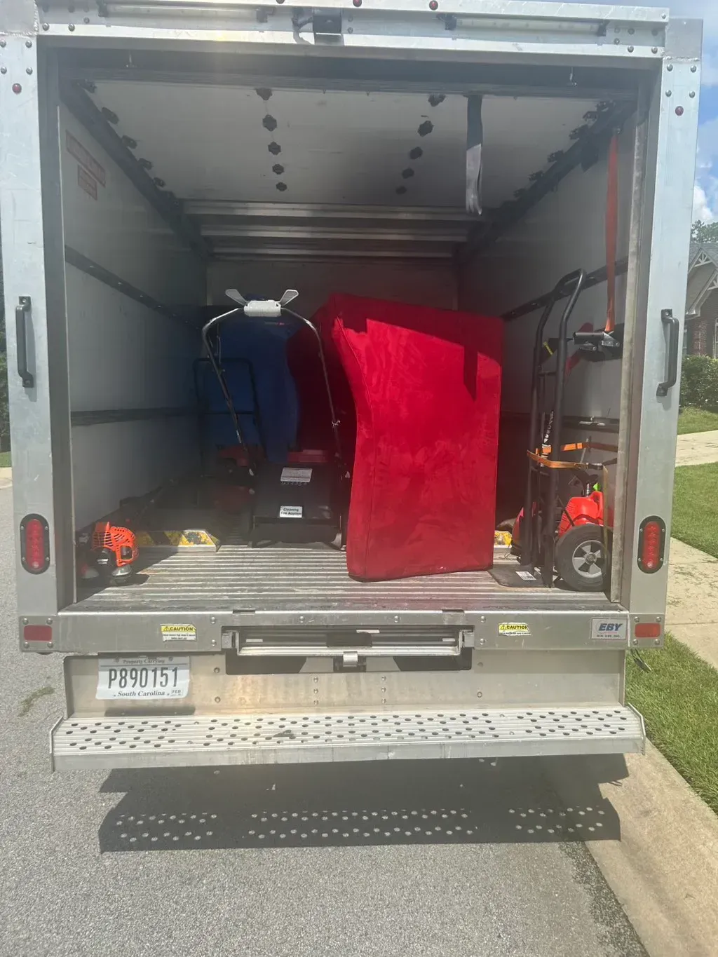 The open rear doors of a parked moving truck reveal a red-covered sofa, a lawnmower, and a dolly inside.