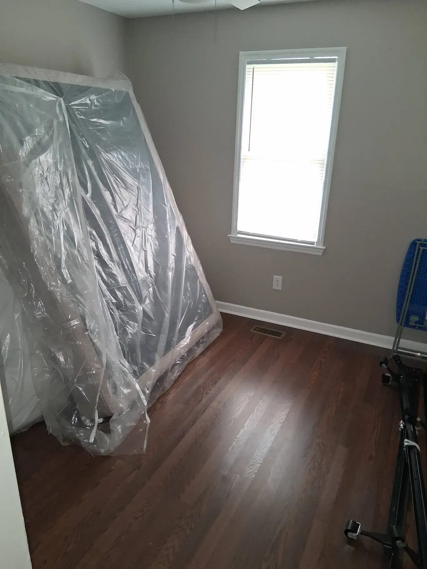 A mattress in a plastic cover leans against a wall in a room with dark wood flooring and a window.