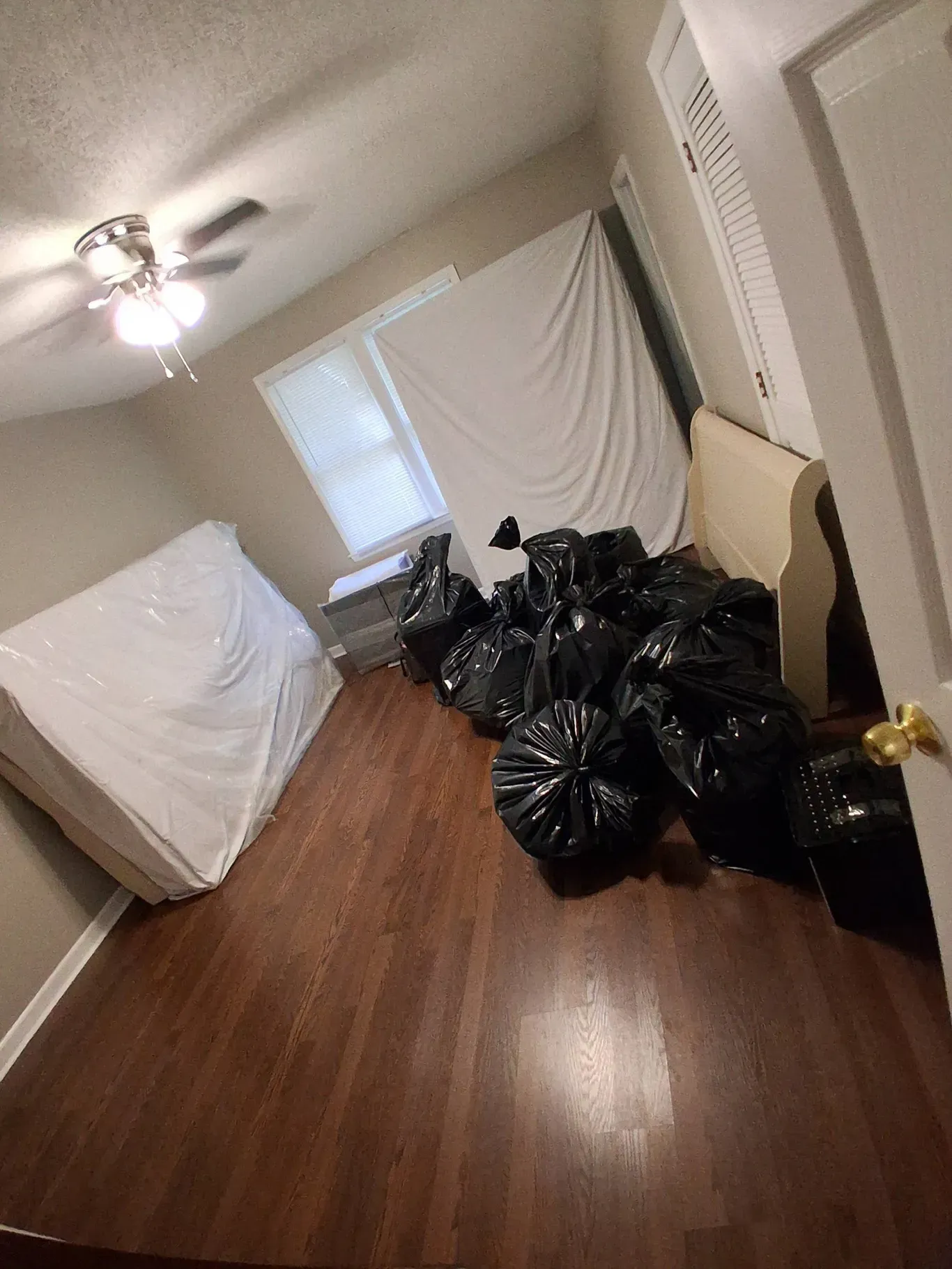 A bedroom with a mattress covered in white fabric, a window, and several black trash bags on the wood floor.