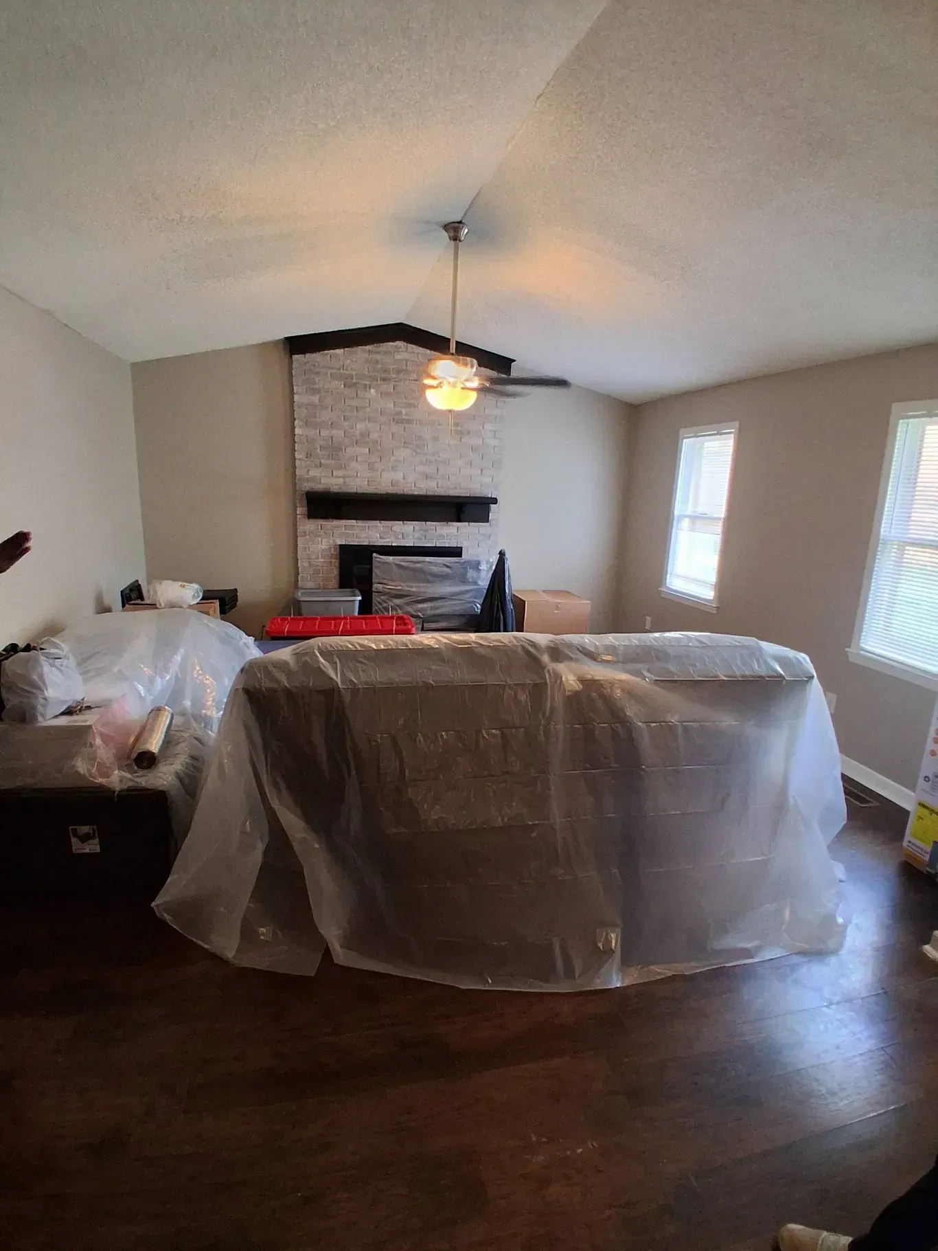 A living room with hardwood floors, a vaulted ceiling, a brick fireplace, and furniture covered in plastic sheeting.