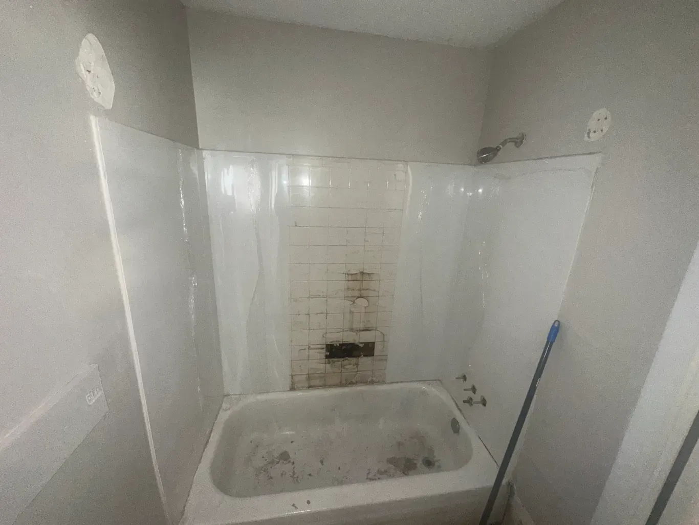 A small, dated bathtub with worn tiled walls, grey painted surround, and a cleaning mop leaning against the side.