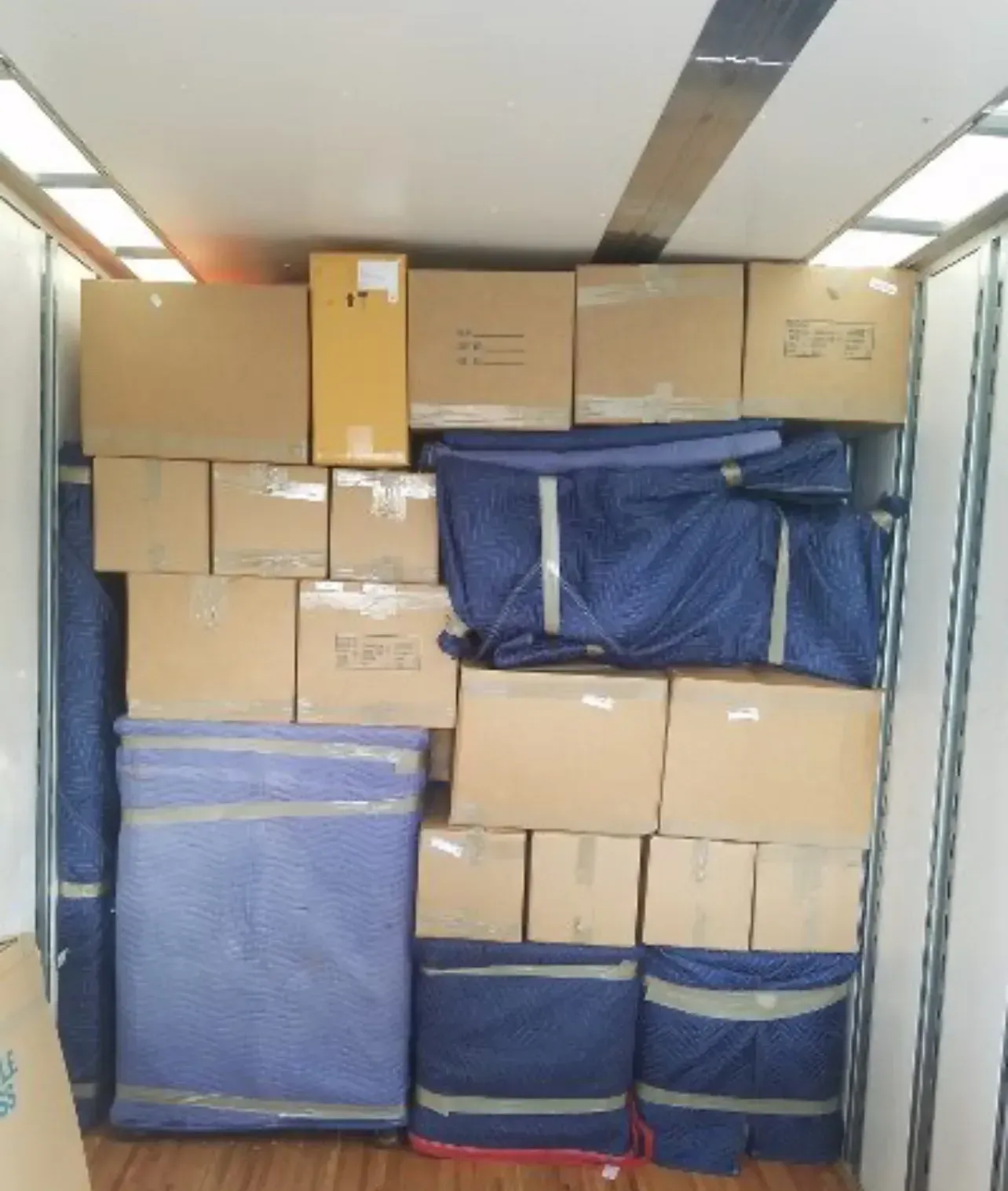A moving truck trailer tightly packed with stacked cardboard boxes and furniture items wrapped in blue protective pads.