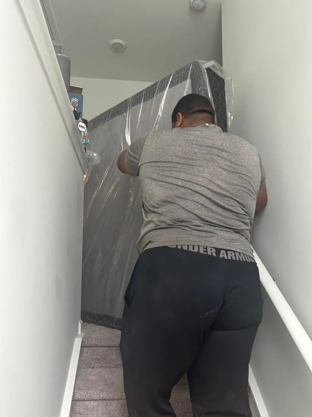 A person in a gray shirt and black athletic pants carefully maneuvers a large, plastic-wrapped mattress up a carpeted stair.