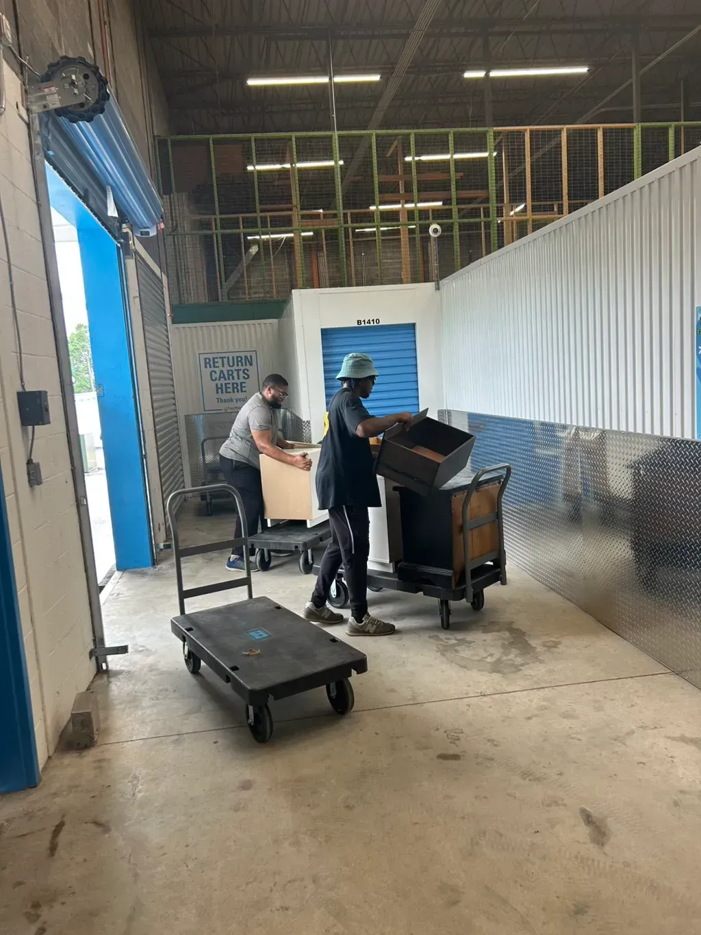 Two people moving boxes in a warehouse area with metal walls and a rolling utility cart.