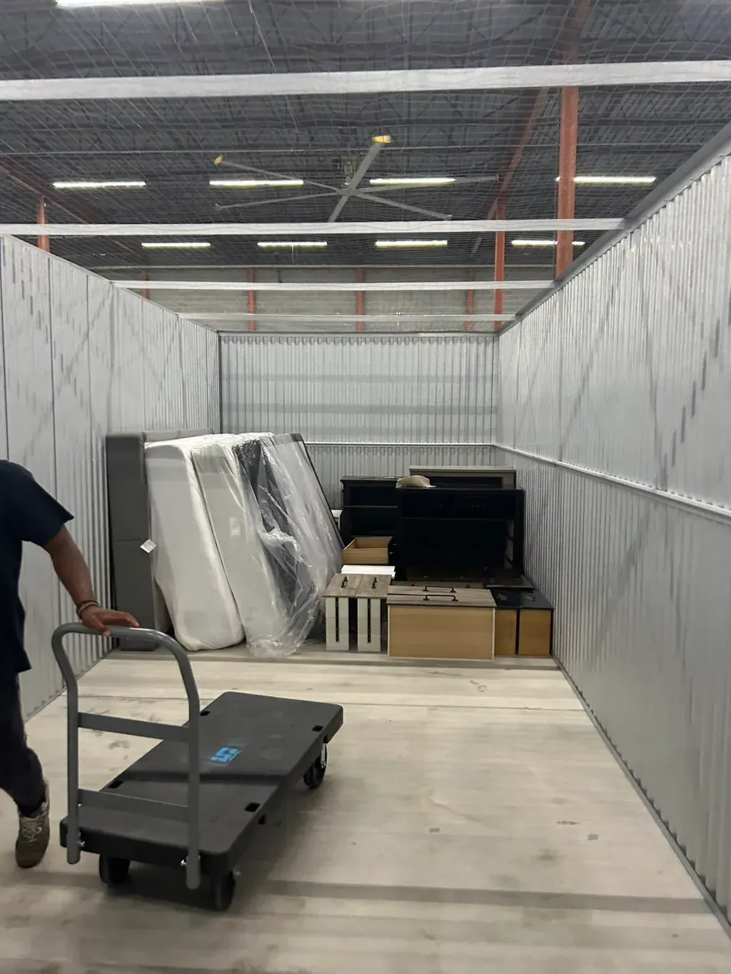A person pushes a flatbed cart into a storage unit containing mattresses, furniture, and cardboard boxes.